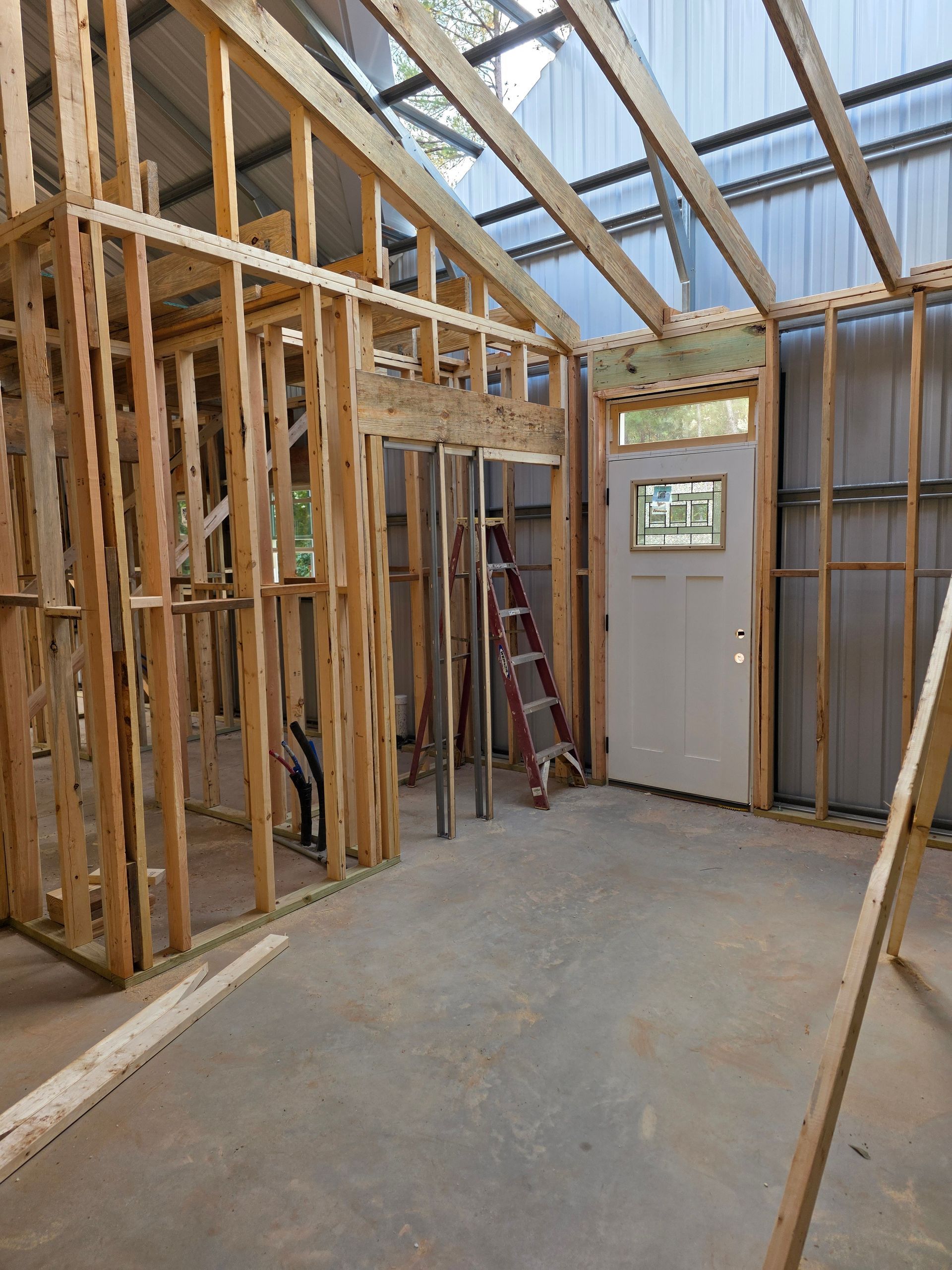 Interior framing of a building under construction, featuring wooden studs, a door, and a ladder.