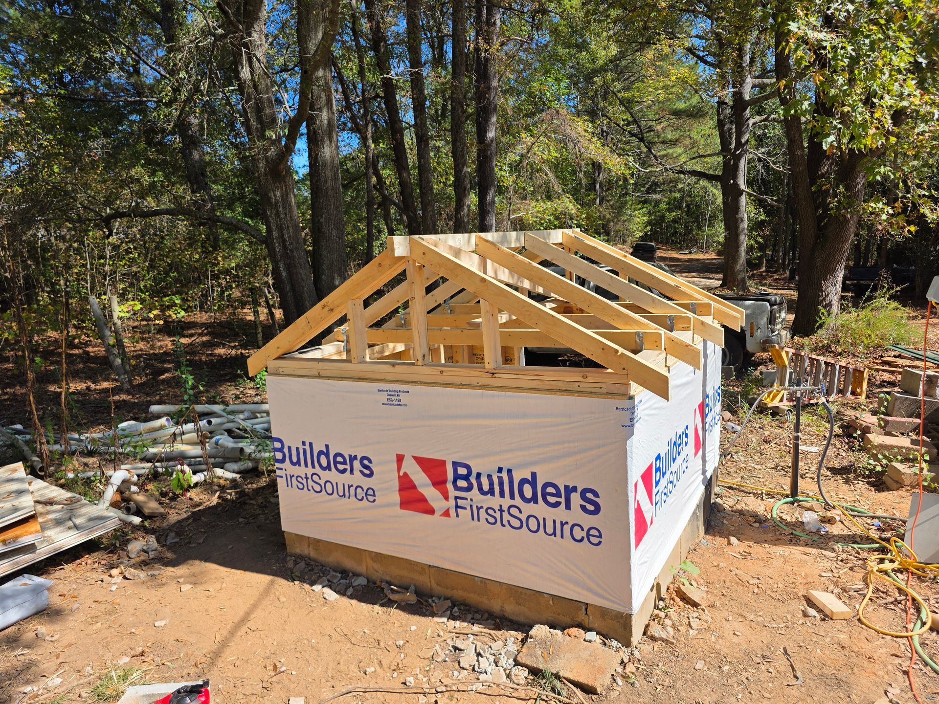 Wooden roof trusses on a small, white-wrapped building in a wooded area.