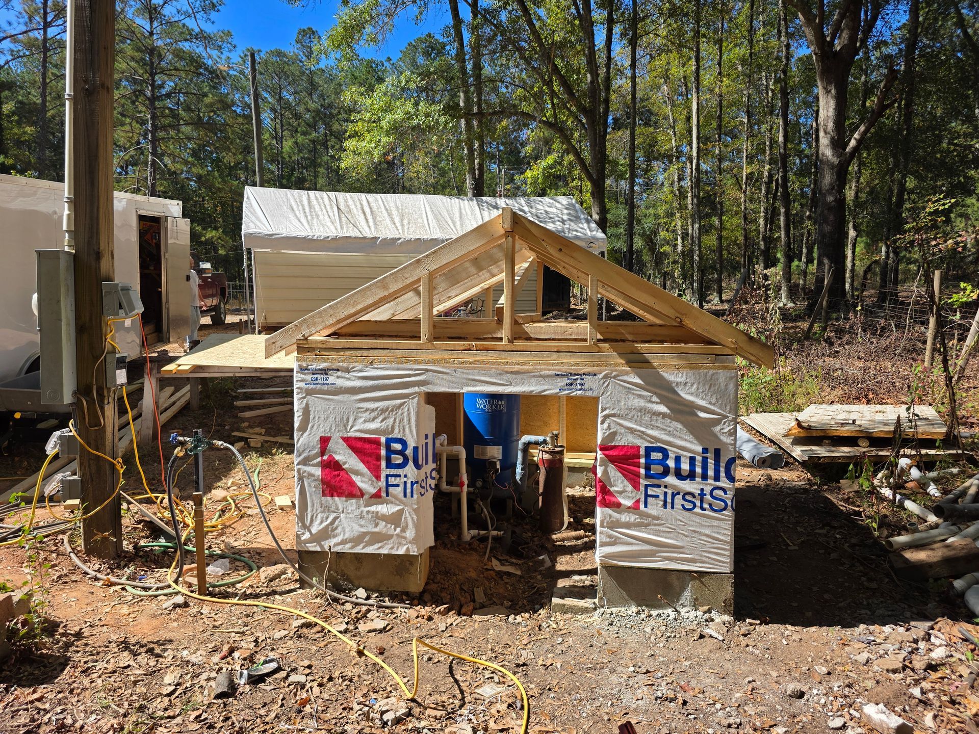 Construction site with a partially built small wooden shed surrounded by trees. Blue and white wrap.