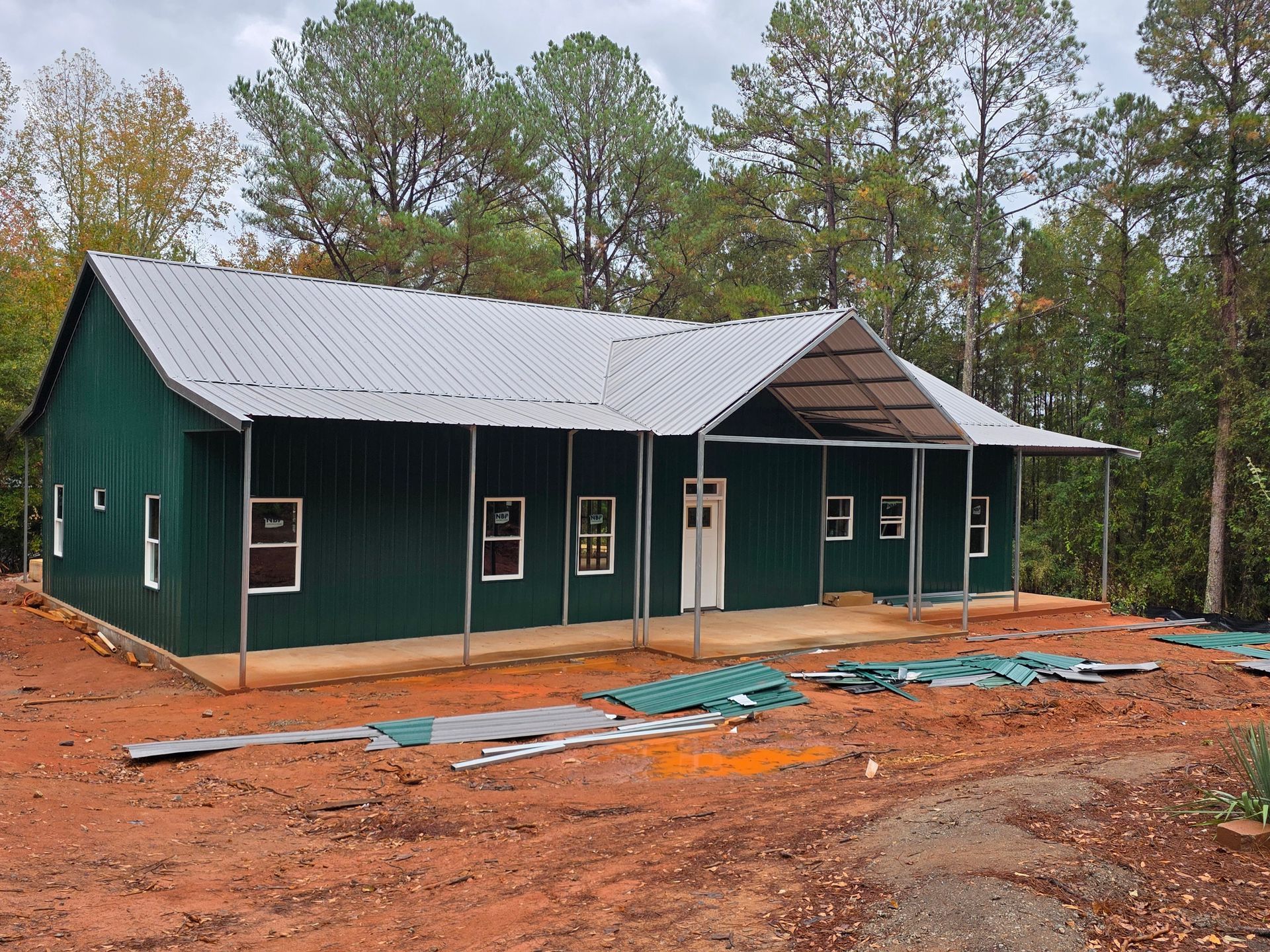 Green metal building with a porch under construction, set in a wooded area.