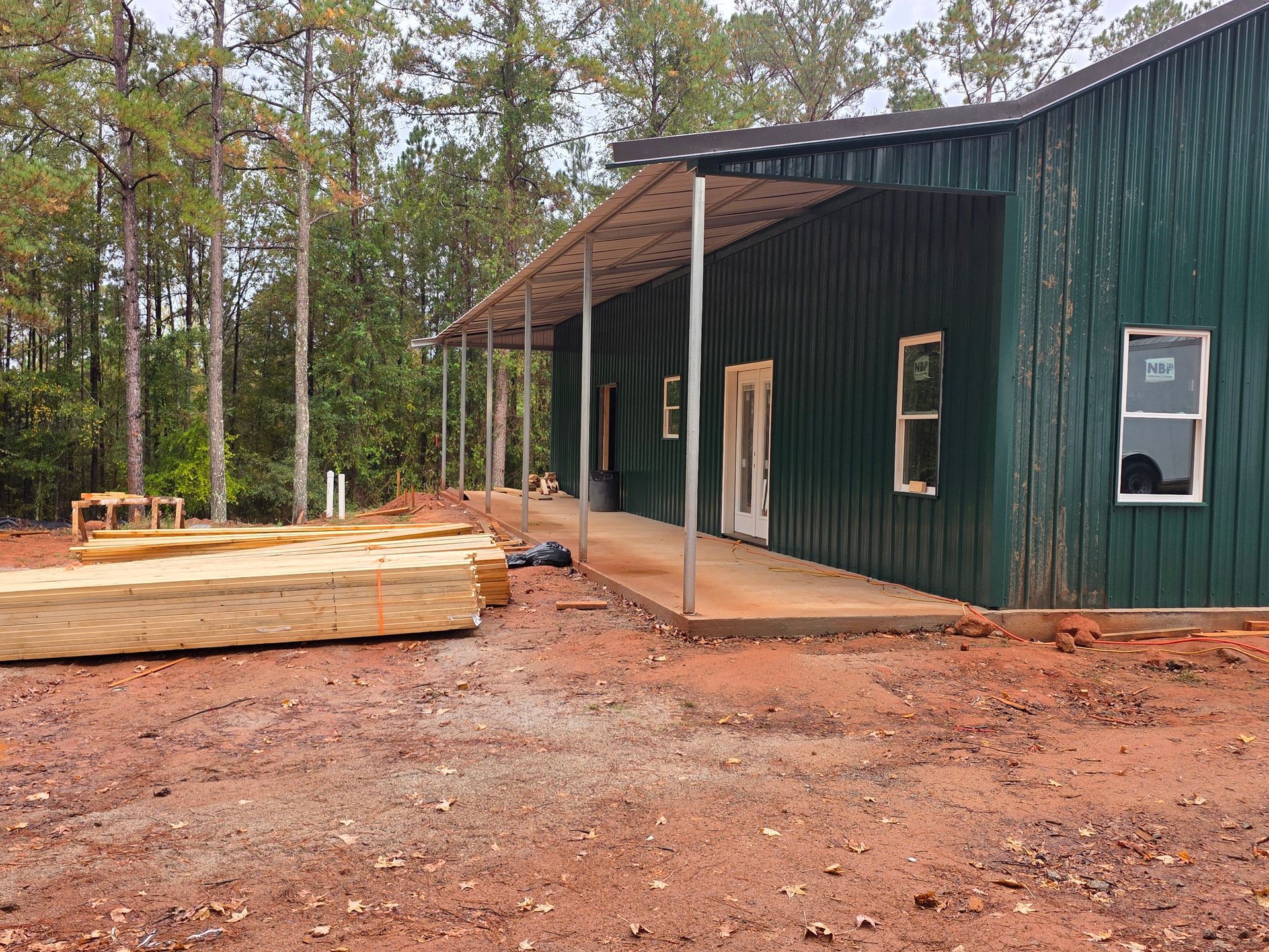 Green-sided building with covered porch under construction on red dirt with lumber stacked to the left.