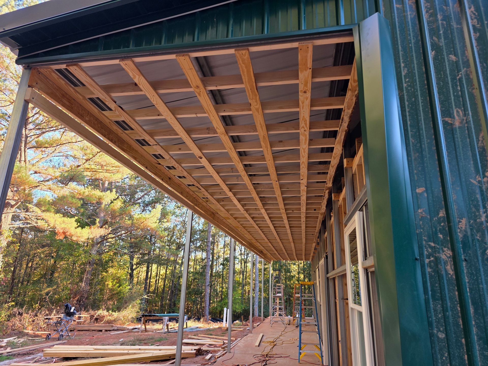 Construction of a porch roof. Wooden frame with metal supports, green metal siding, and a forest background.
