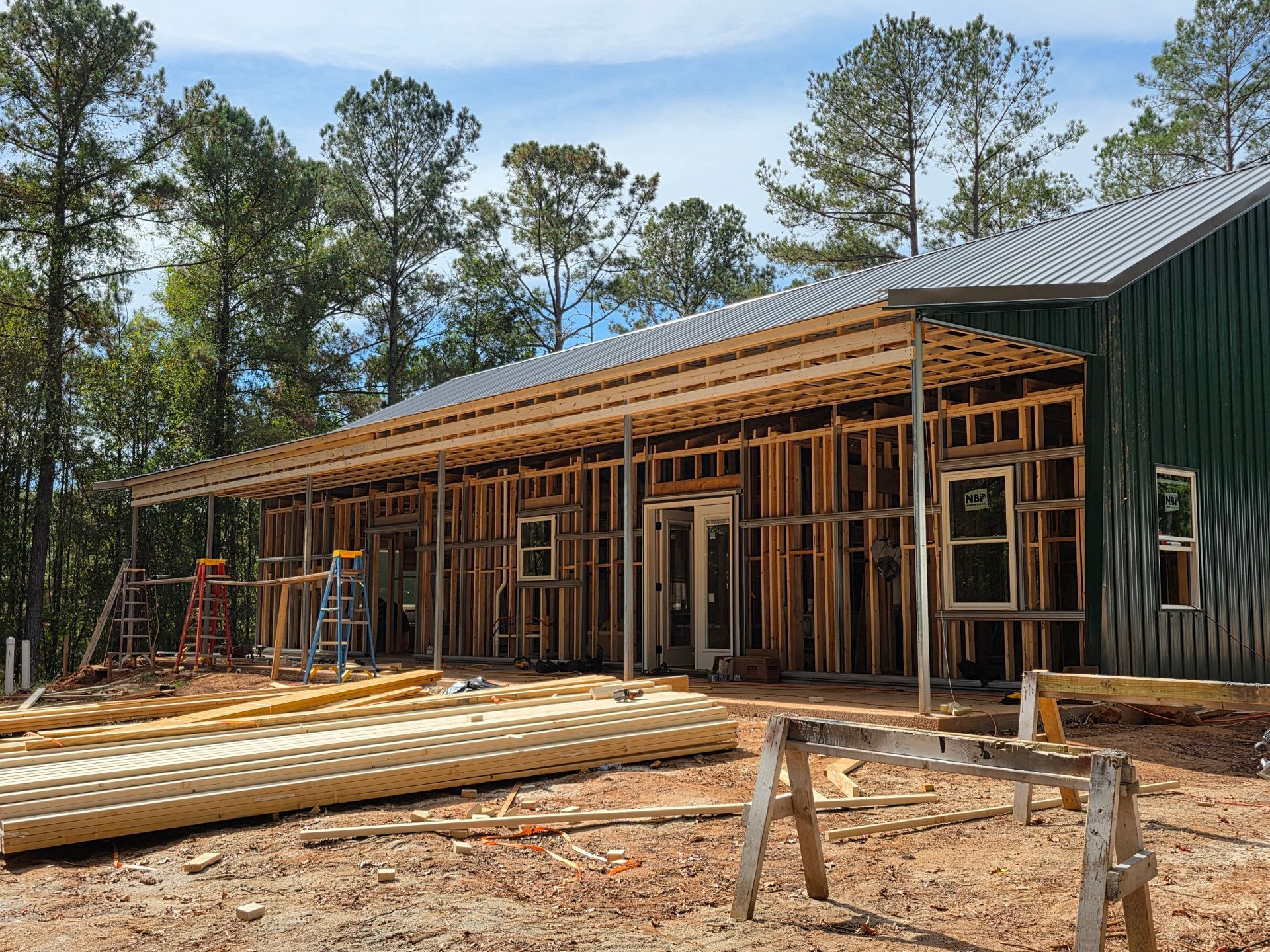 A building under construction with exposed wooden framing and a green metal exterior, porch roof.