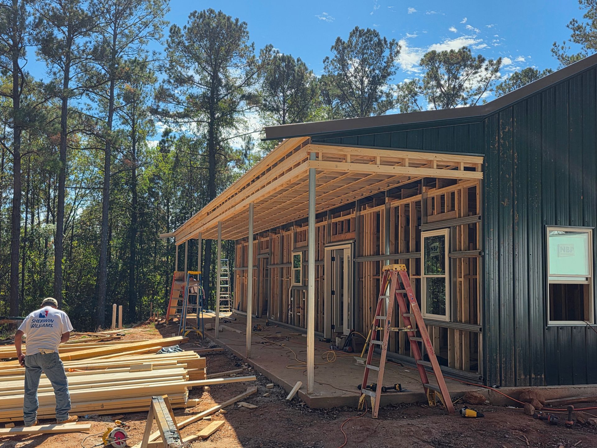 Construction of a green metal building with a wooden porch; a worker stands by lumber in a wooded area.