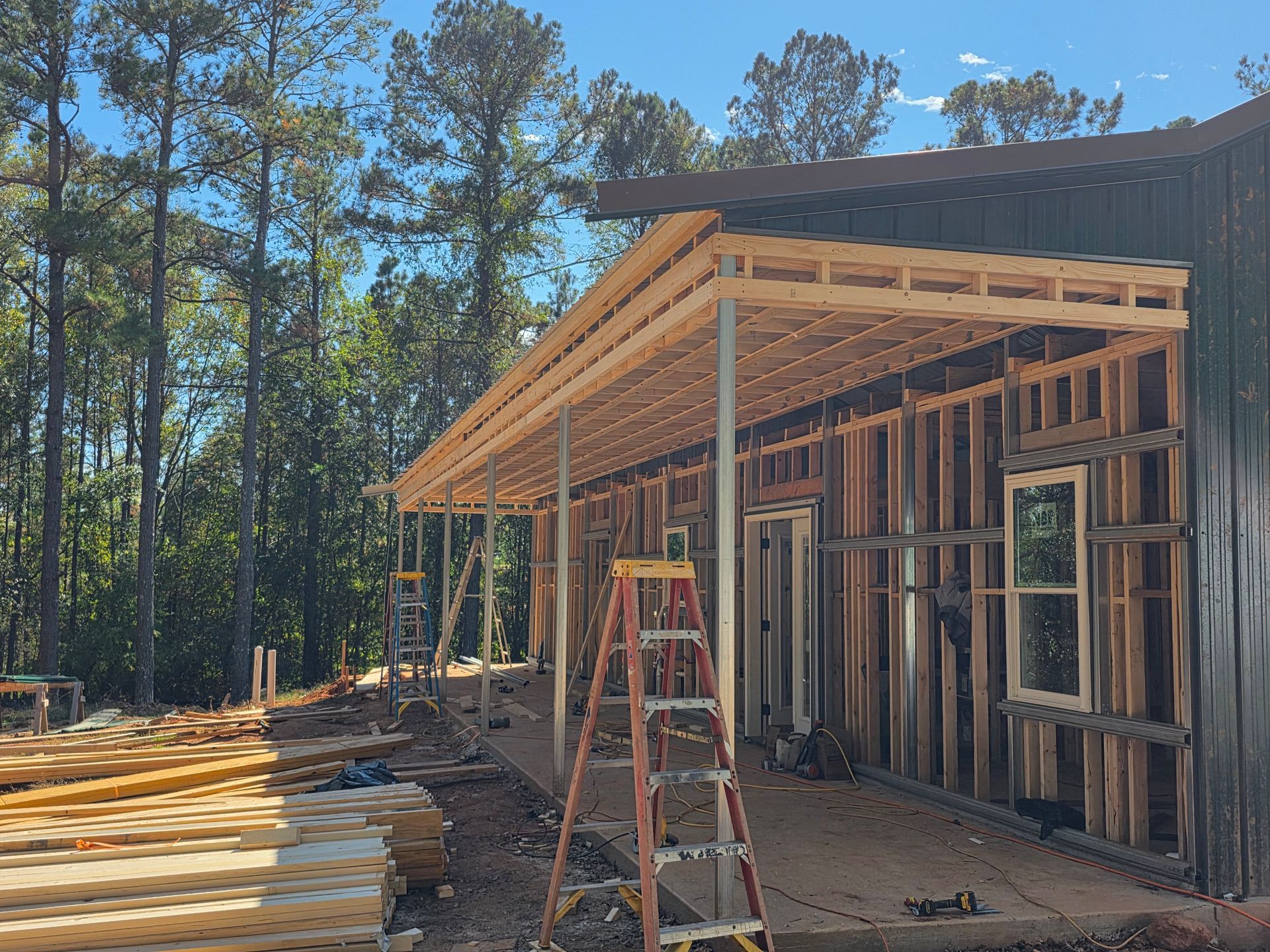 Construction of a building with a wooden framework, porch roof, and ladder outdoors; trees in the background.