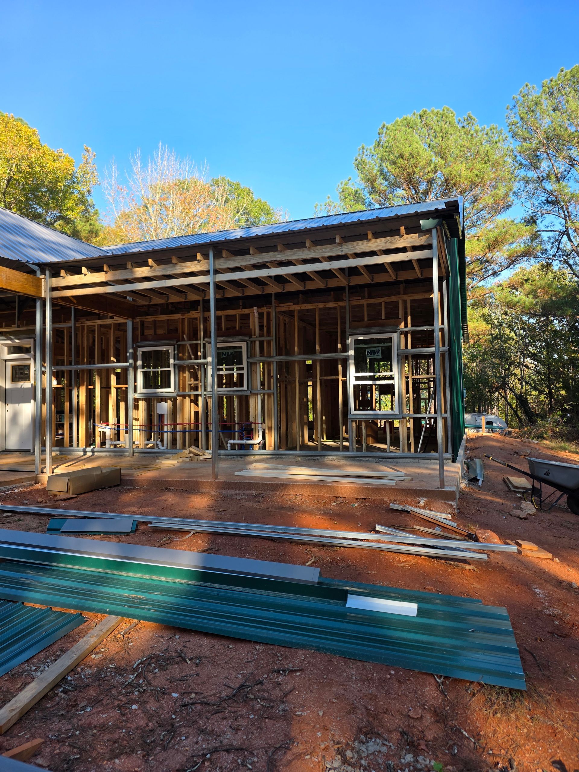 Framed wooden structure of a building under construction, with metal roofing, windows, and green corrugated sheets.