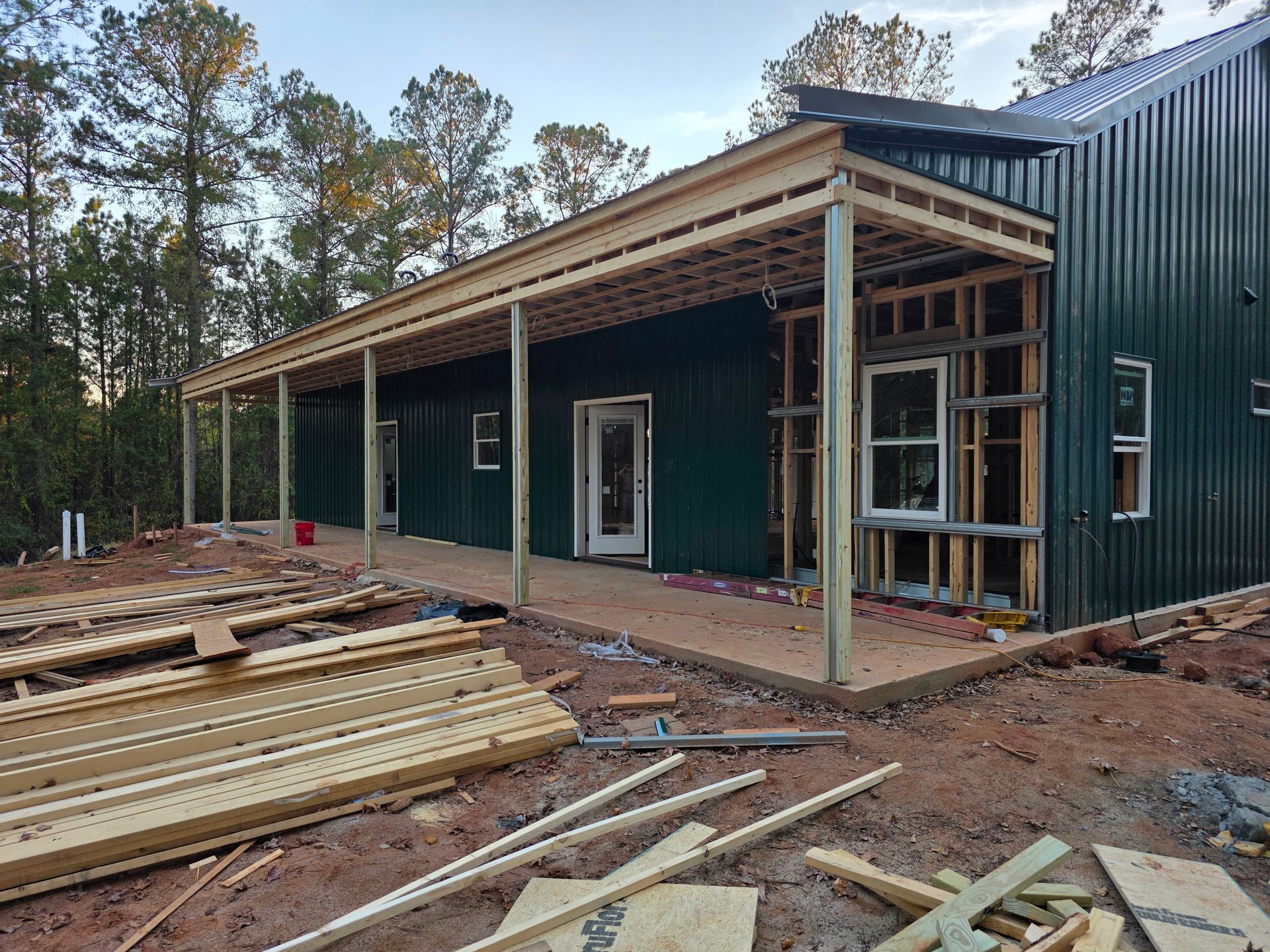 A building under construction with a wooden porch attached to the side. Green siding and a wooded background.