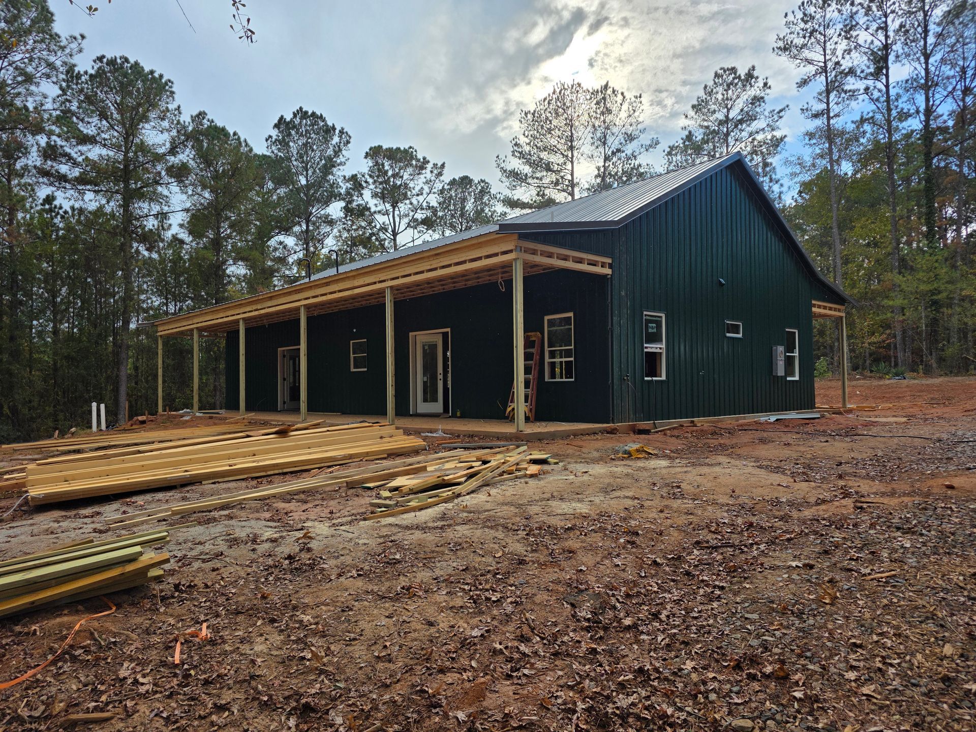 Green metal-clad building under construction with a wraparound porch in a wooded area.