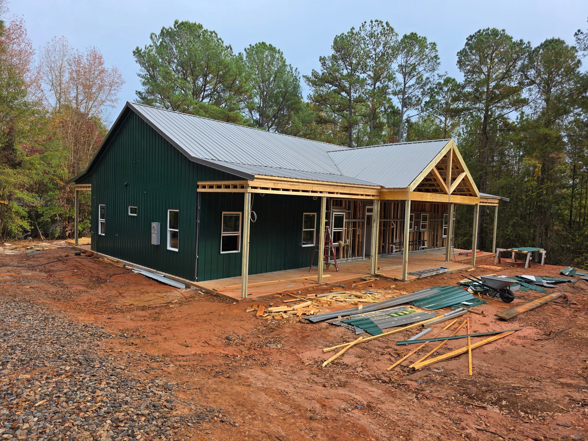 Green metal-clad building under construction; wooden porch and roof framing. Gray roof, brown dirt ground.