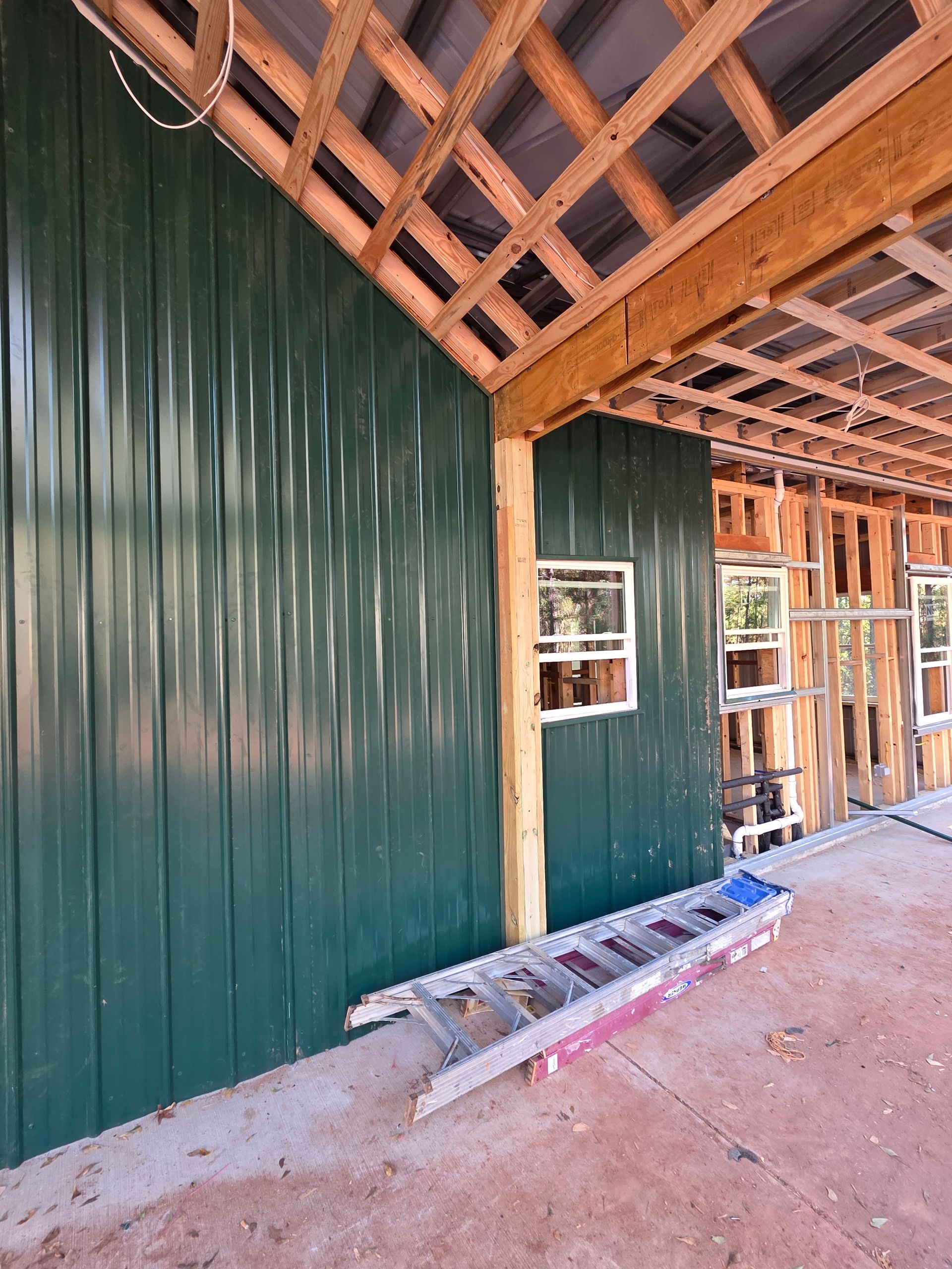 Green metal siding on a building under construction, exposed wooden beams, ladder, windows.