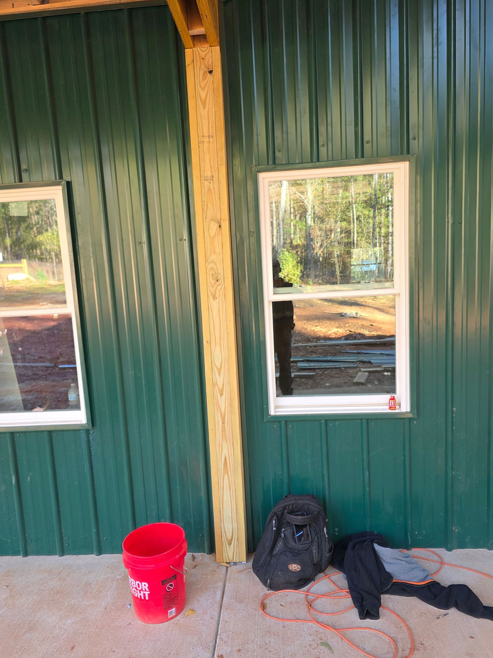 Exterior of a green metal building with windows, a wooden support beam, and construction materials on the concrete slab.
