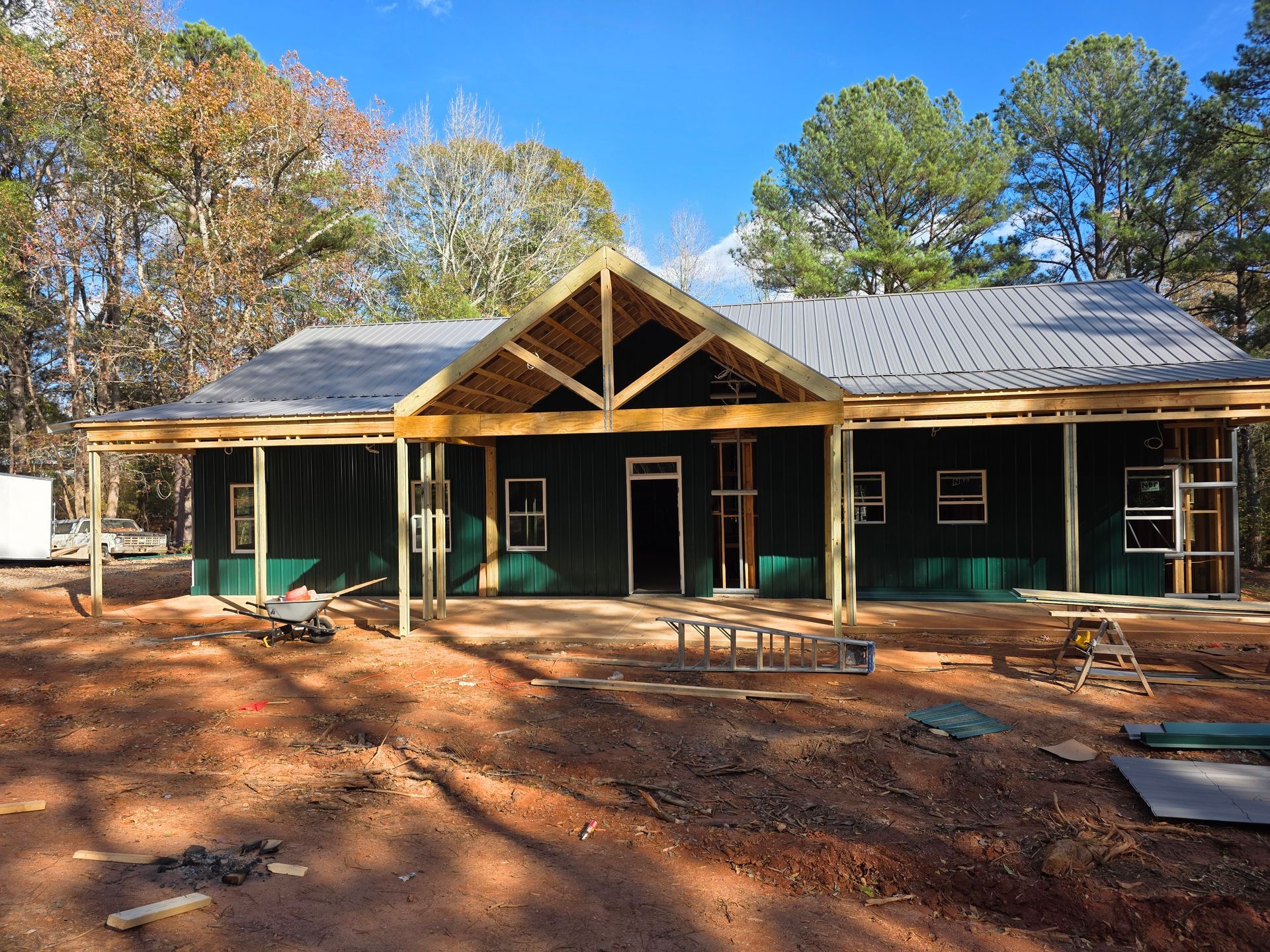 House under construction with green siding, porch, and a partially built roof in a wooded area.