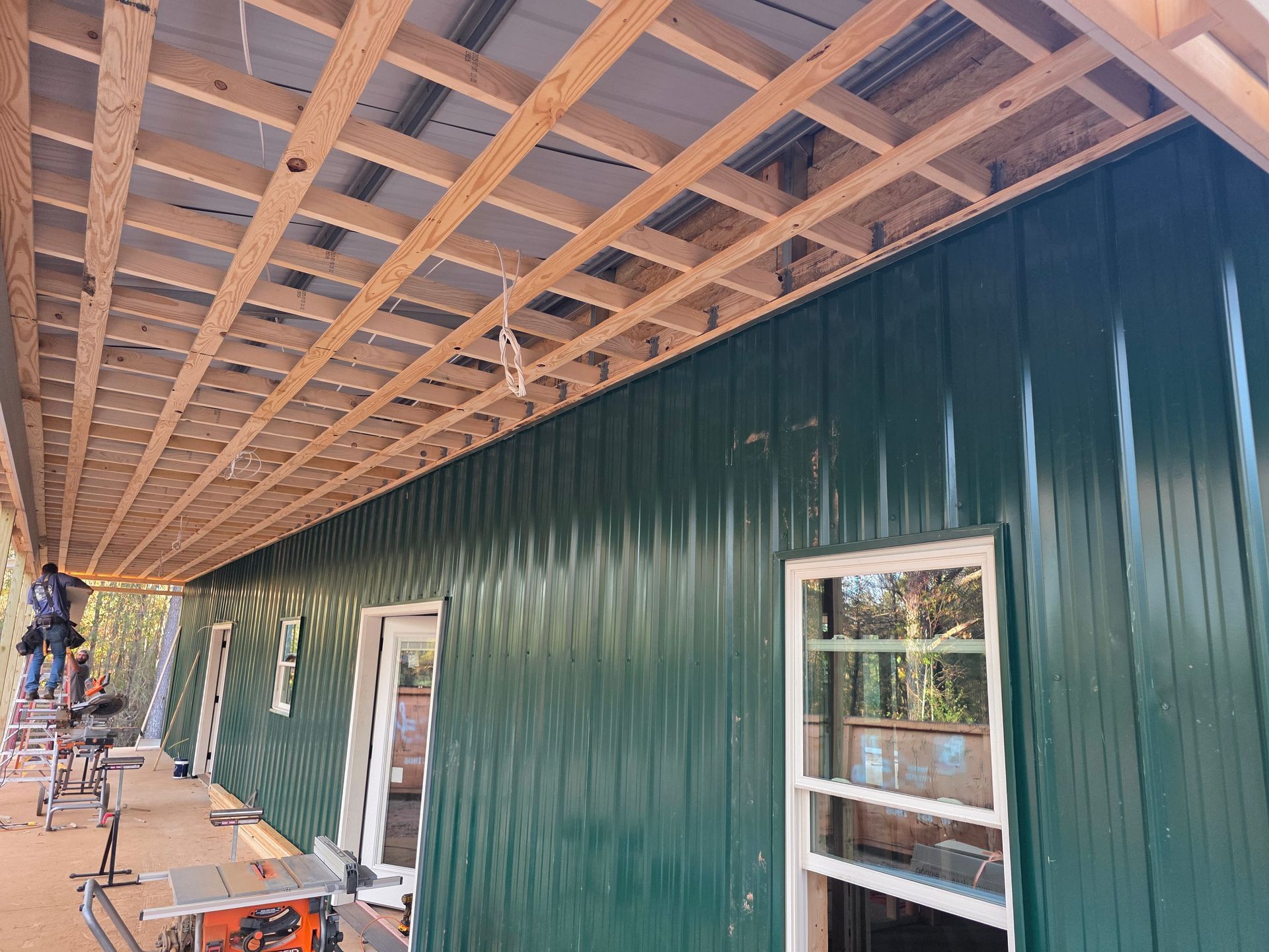 Construction of a building with green siding and exposed wood beams. A window and door are visible.