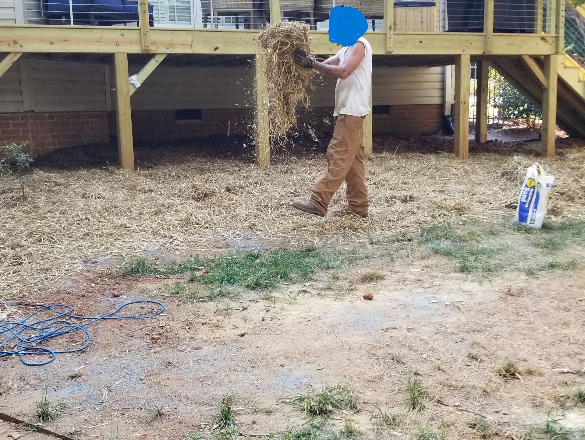 Person spreading mulch under a wooden deck. The person wears brown pants, a white shirt, and gloves.