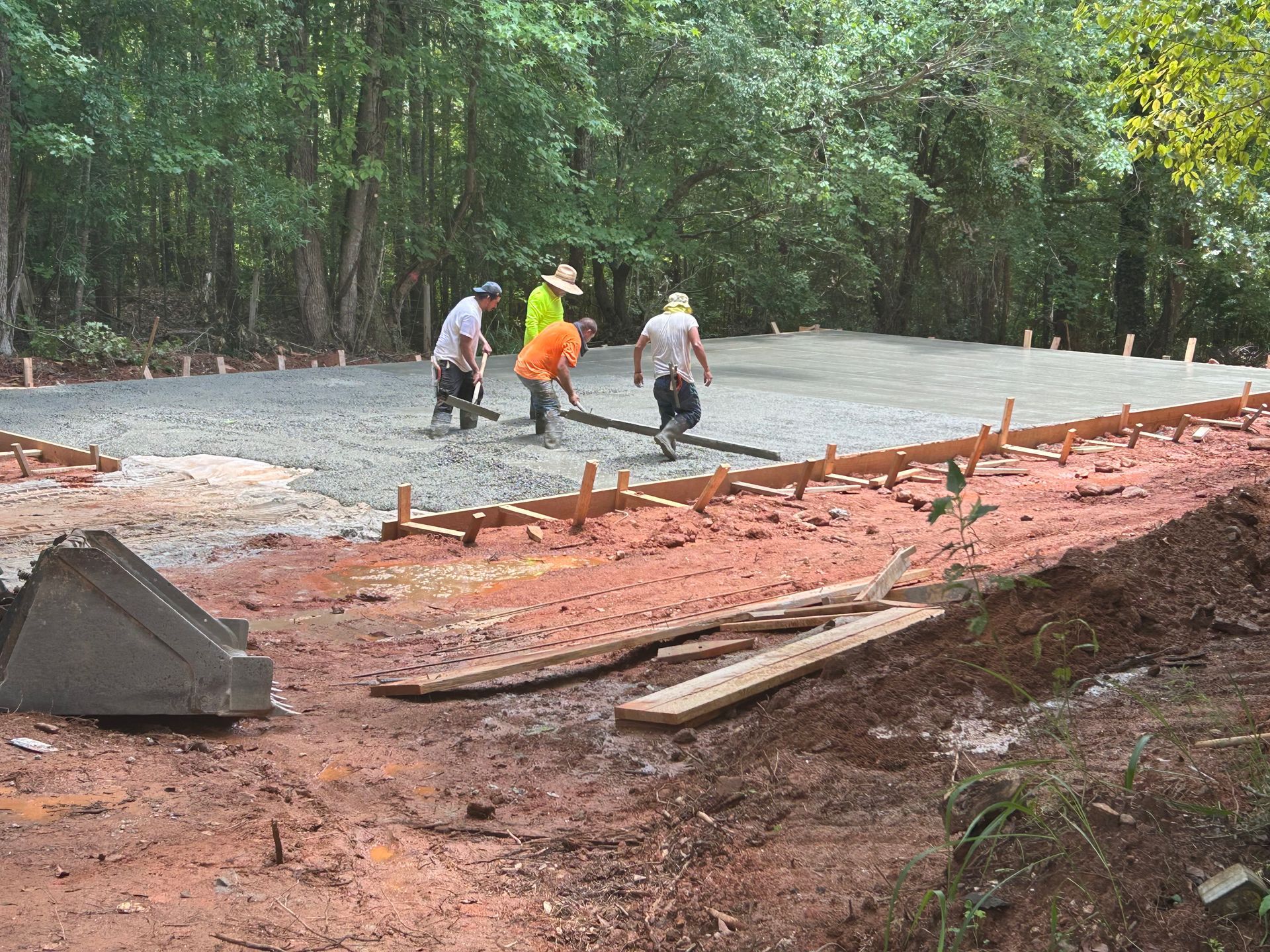 Construction workers smoothing wet concrete for a foundation, outdoors in a wooded area.