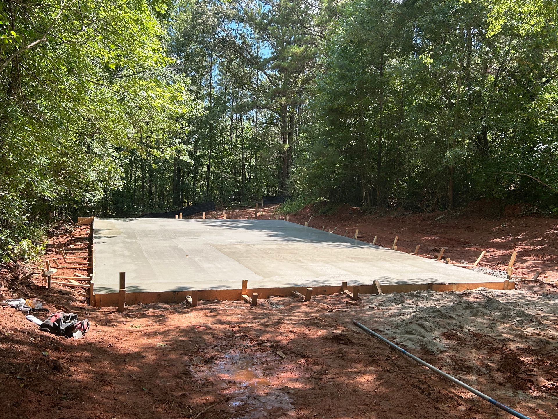 A freshly poured concrete slab in a wooded area, surrounded by wooden framing and red soil.