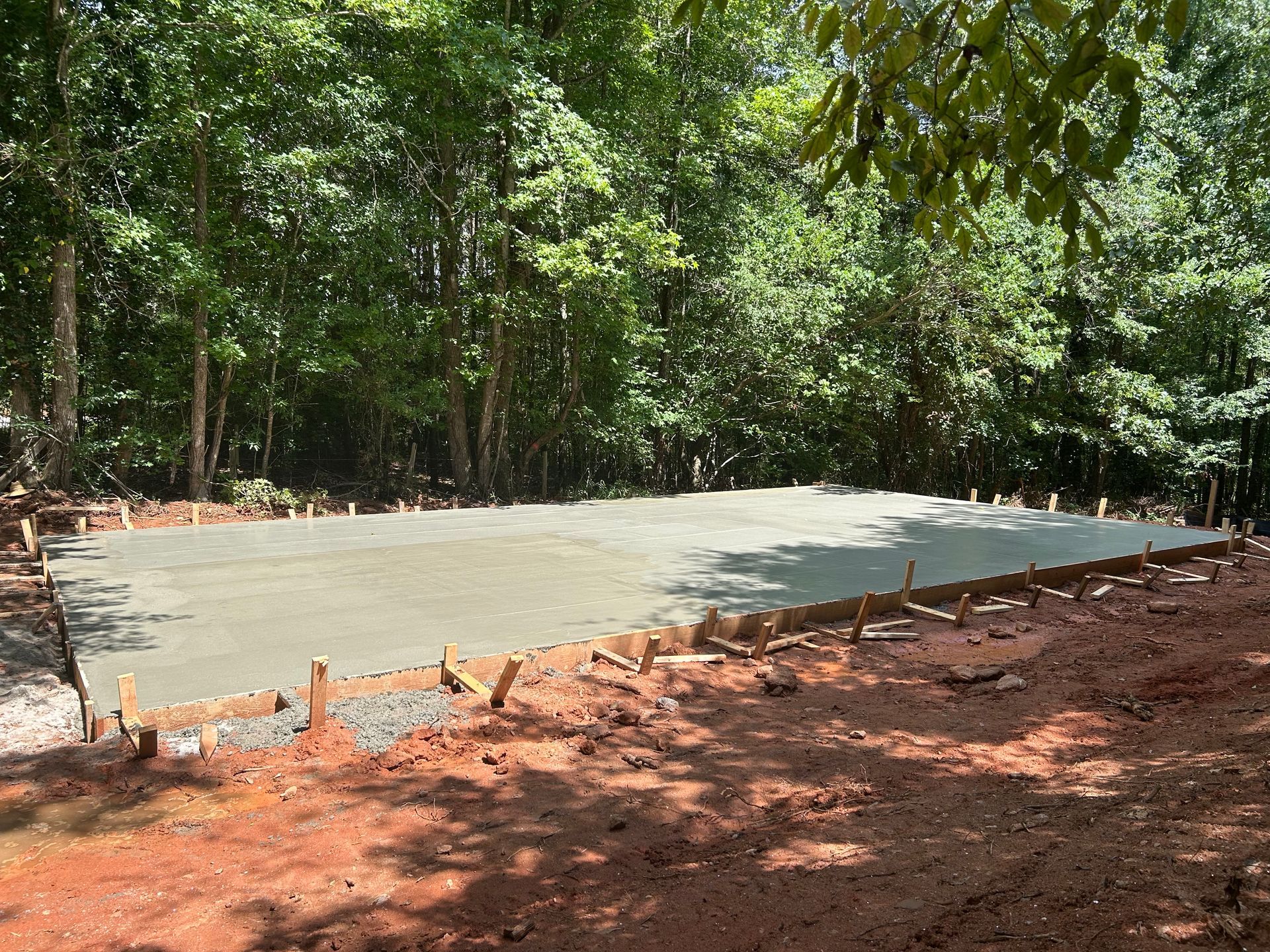 Freshly poured concrete slab in a wooded area, surrounded by wooden forms and red soil.