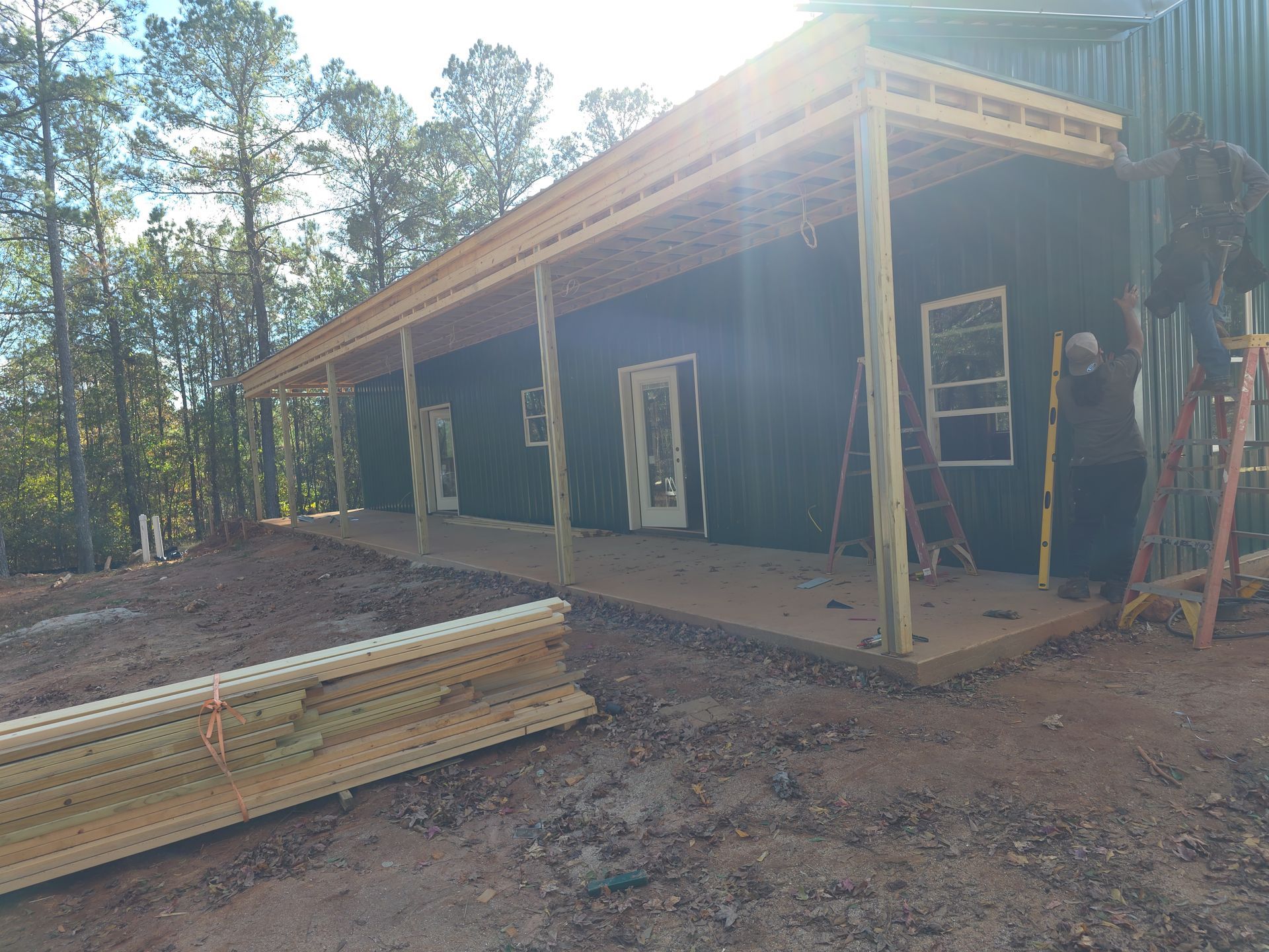 Construction of a house with a covered porch. Green exterior, wooden supports and roof, ladders, and lumber on the ground.