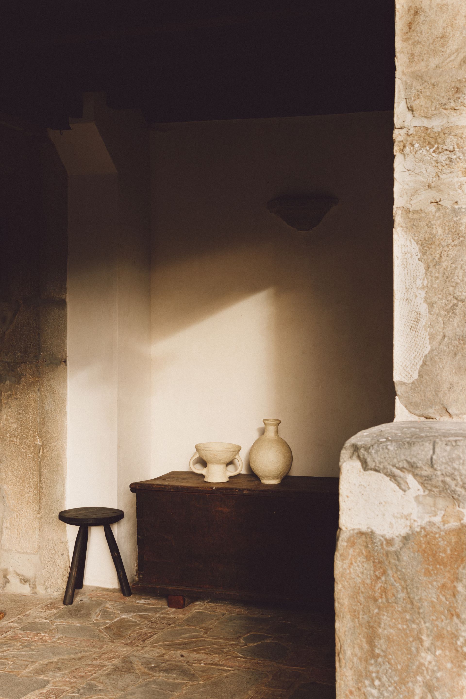 Stone alcove with dark wooden chest, small stool, and pottery, lit by sunlight.