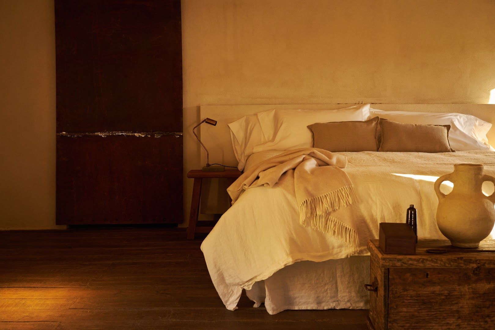 Bedroom with white bedding, wooden floors, and a dark brown, abstract wall piece.