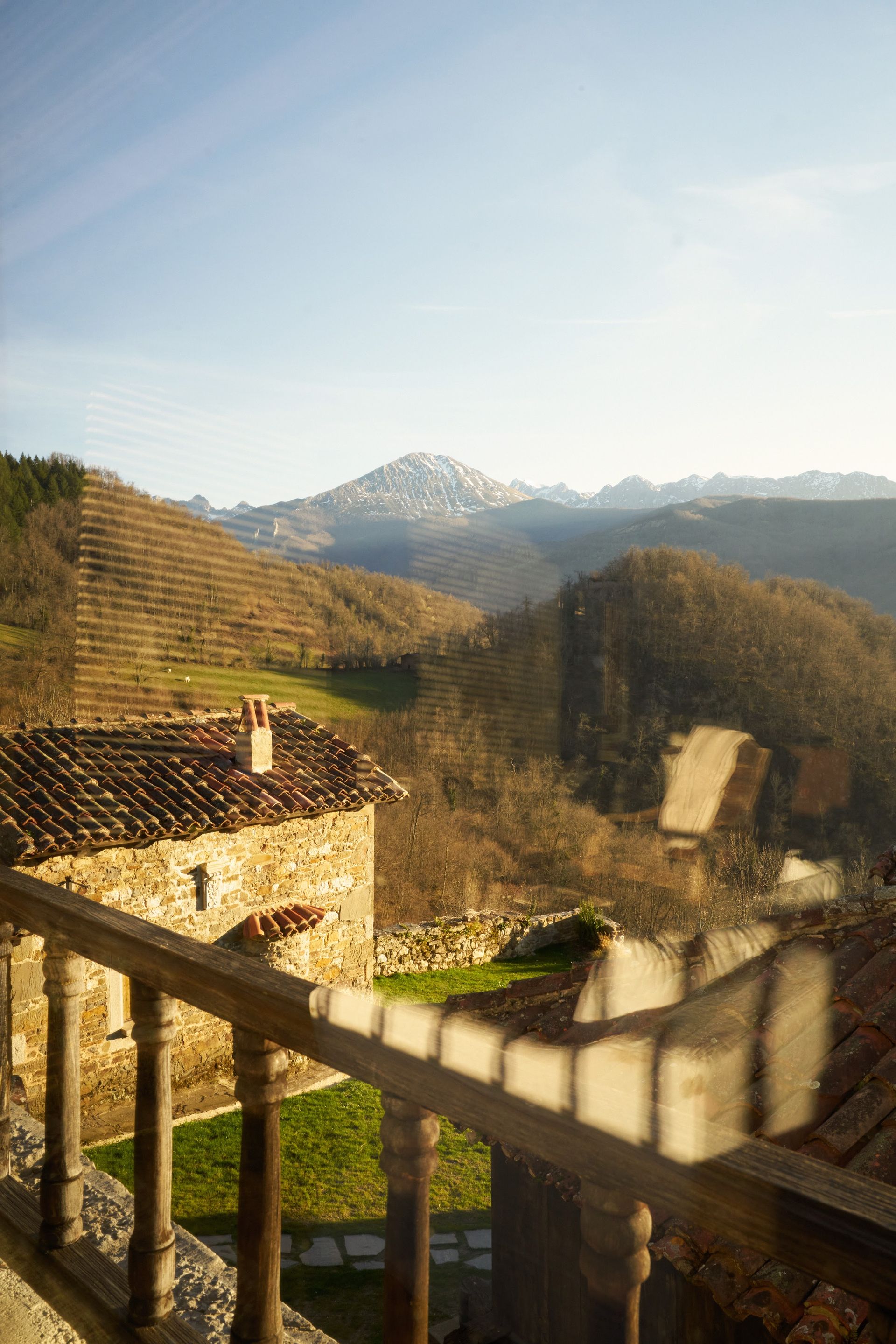 Stone building with red-tiled roof, wooden railing, and mountain landscape under a blue sky.