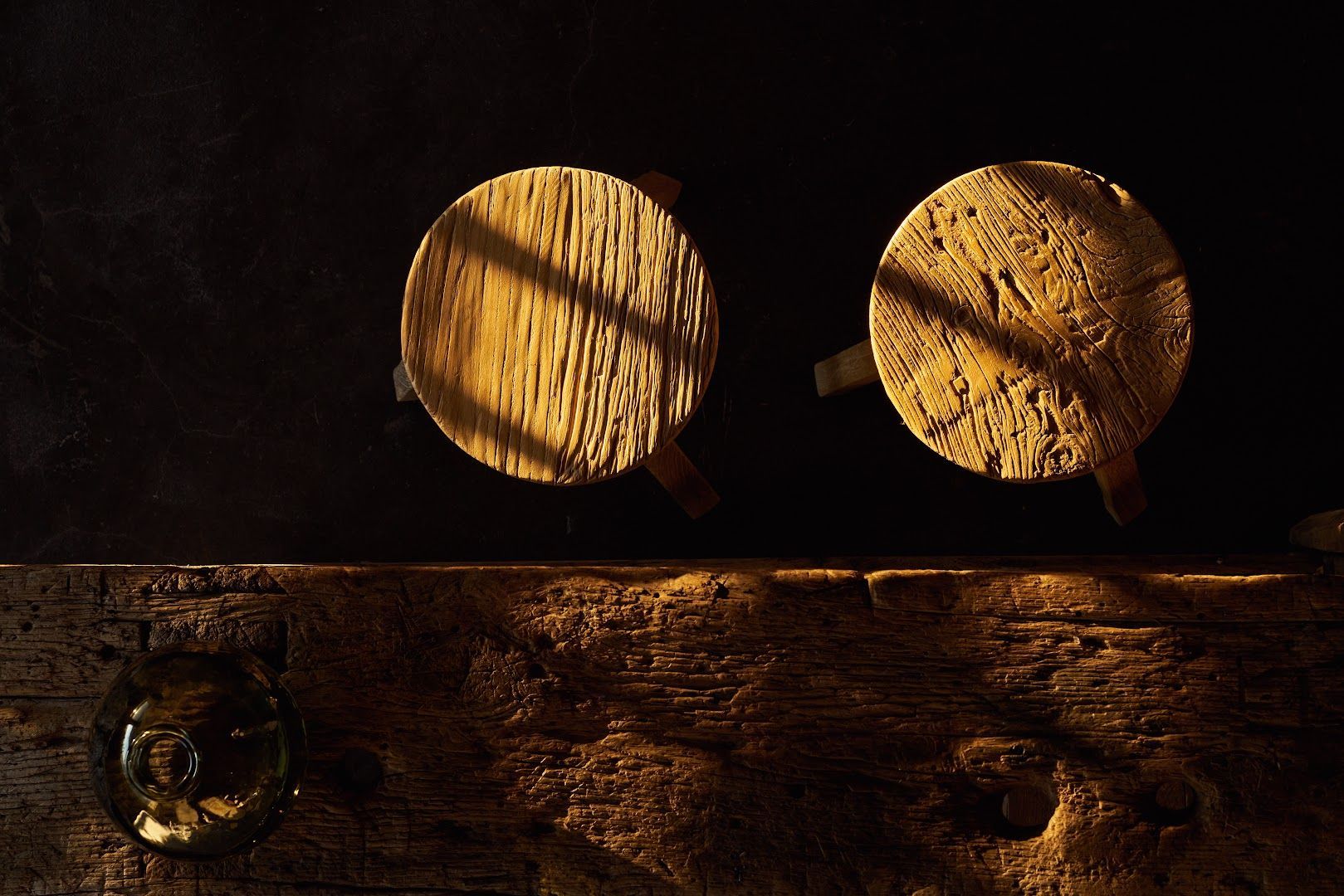 Two wooden stools, lit by sunlight, casting shadows on a wooden beam.