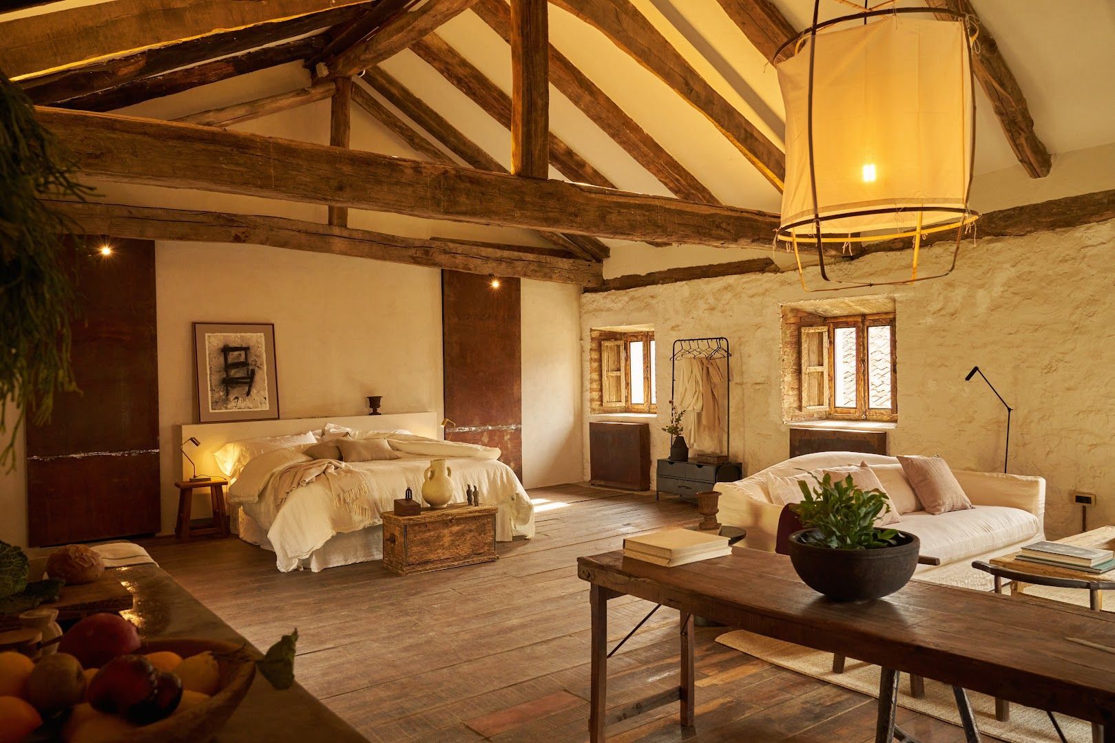 Spacious bedroom with wooden beams, stone walls, and a large bed. A sofa and table are present.
