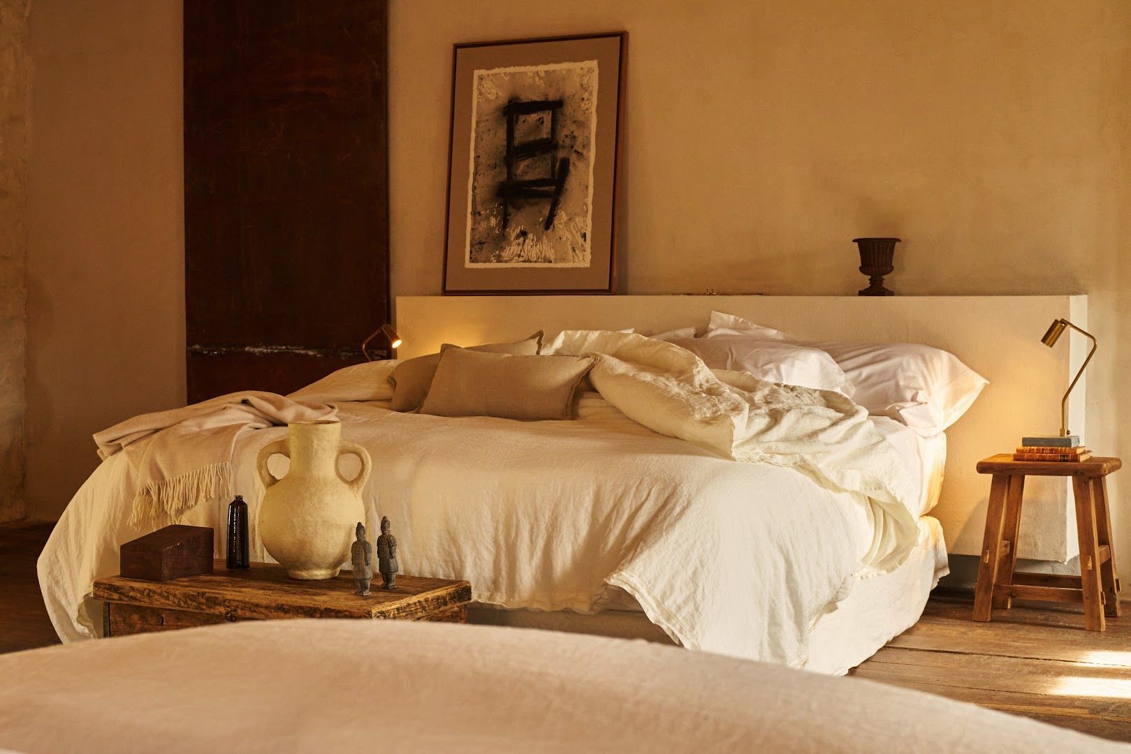 Bedroom with unmade white bed, artwork, small table, and neutral color palette.