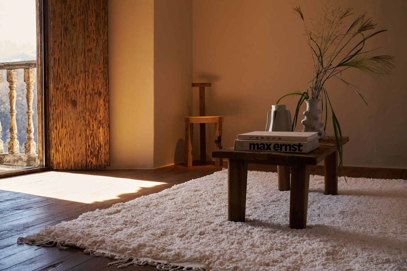 Cozy living room corner with white rug, wooden table with books, vase with plants, and a balcony.