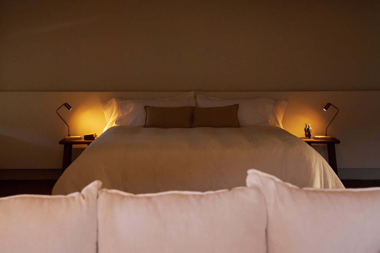 Cozy bedroom with large bed, lit by bedside lamps and soft overhead light. Beige tones, pillows, and a couch in foreground.