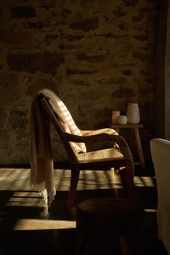 Wooden armchair with blanket, lit by sunlight, against a stone wall.