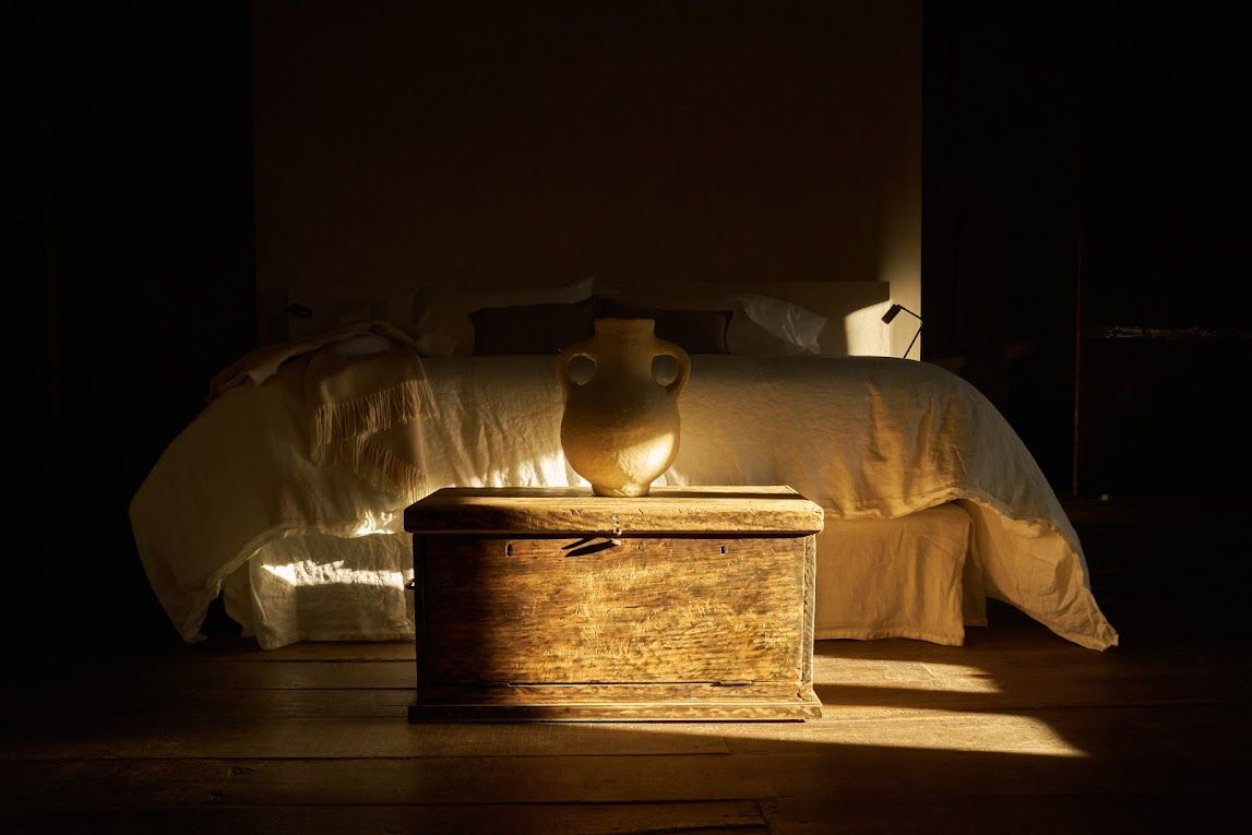 Wooden chest in sunlight, topped with a vase, in front of a bed. Dark, moody interior.