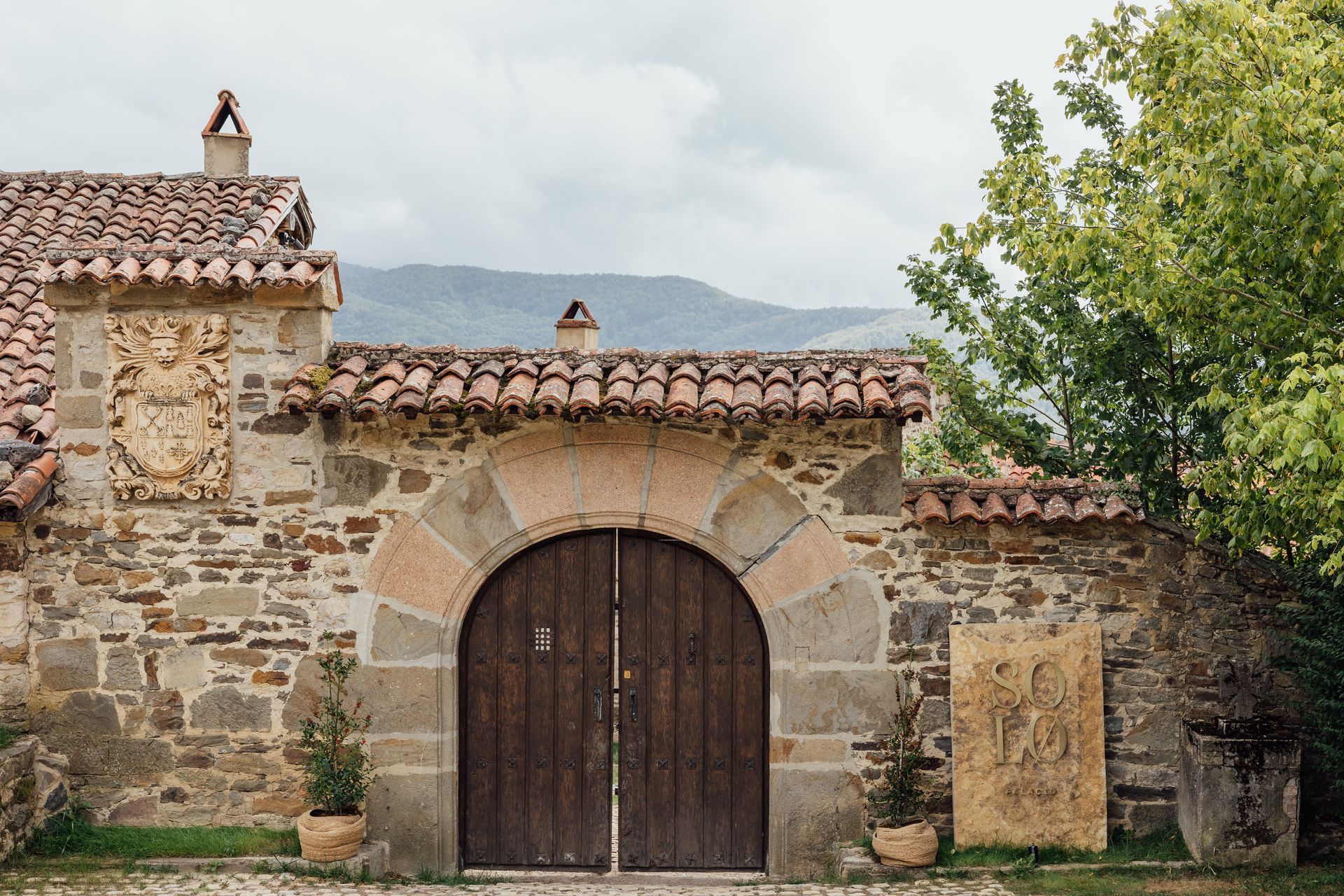Stone wall with arched wooden doors, red-tiled roof, and a decorative stone emblem under a cloudy sky.