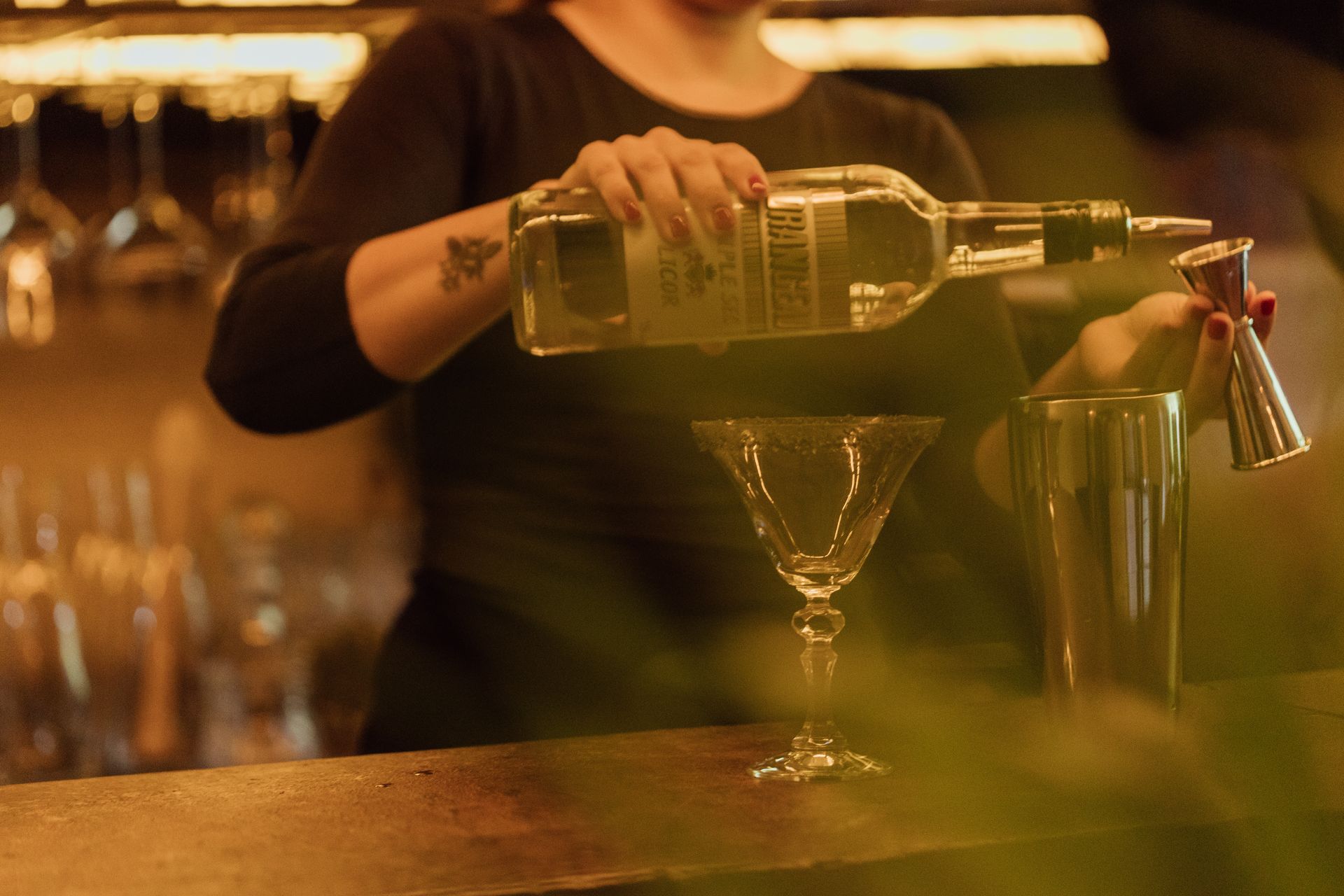 Bartender pouring liquor into a martini glass, next to a shaker, at a bar.