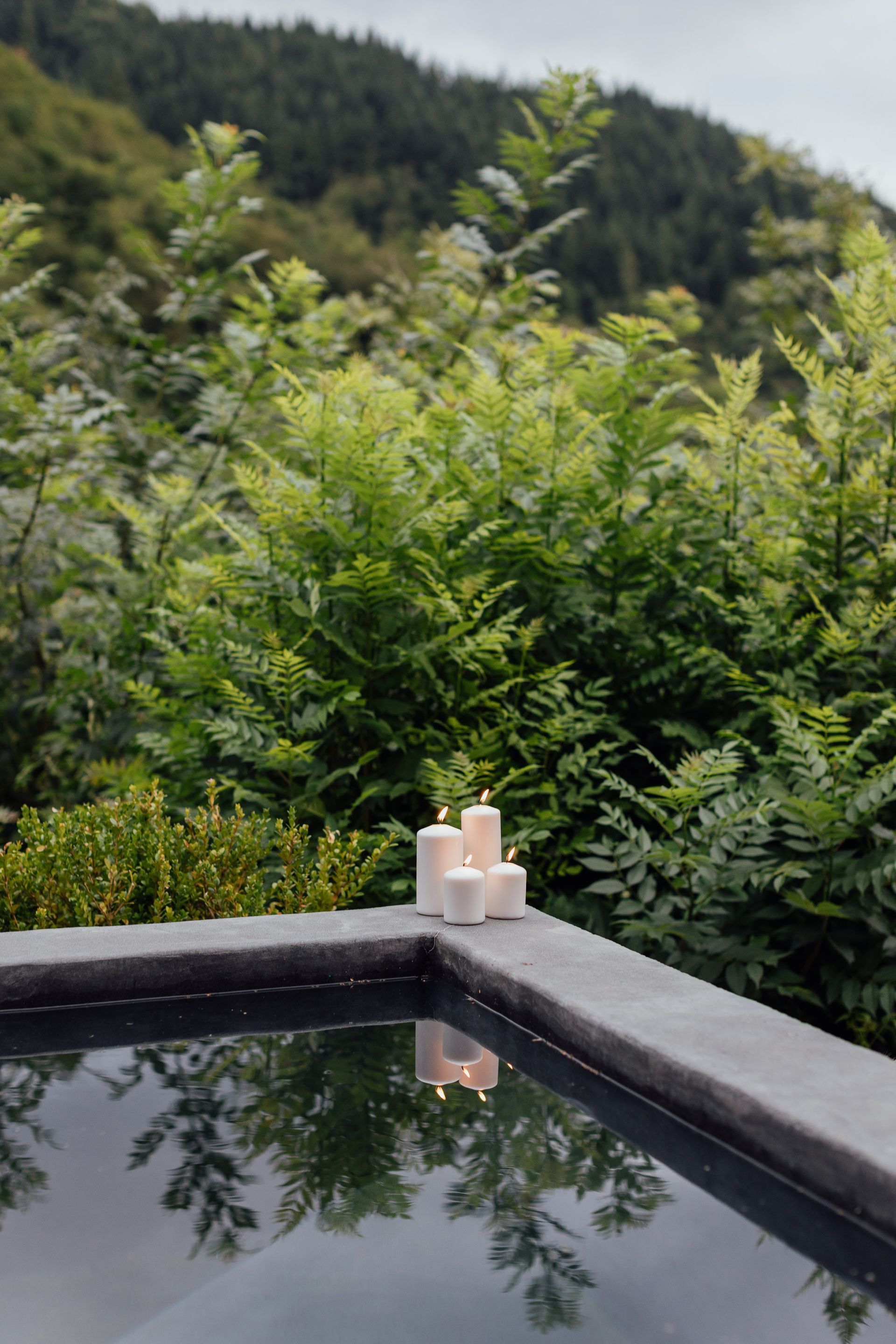 Lit candles on the edge of a pool, reflecting in the water, with a backdrop of green trees and a cloudy sky.