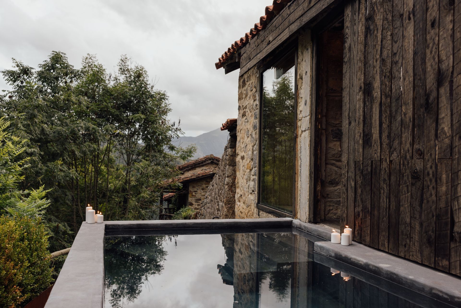Outdoor pool next to a rustic cabin with large window reflecting cloudy sky and surrounding trees.