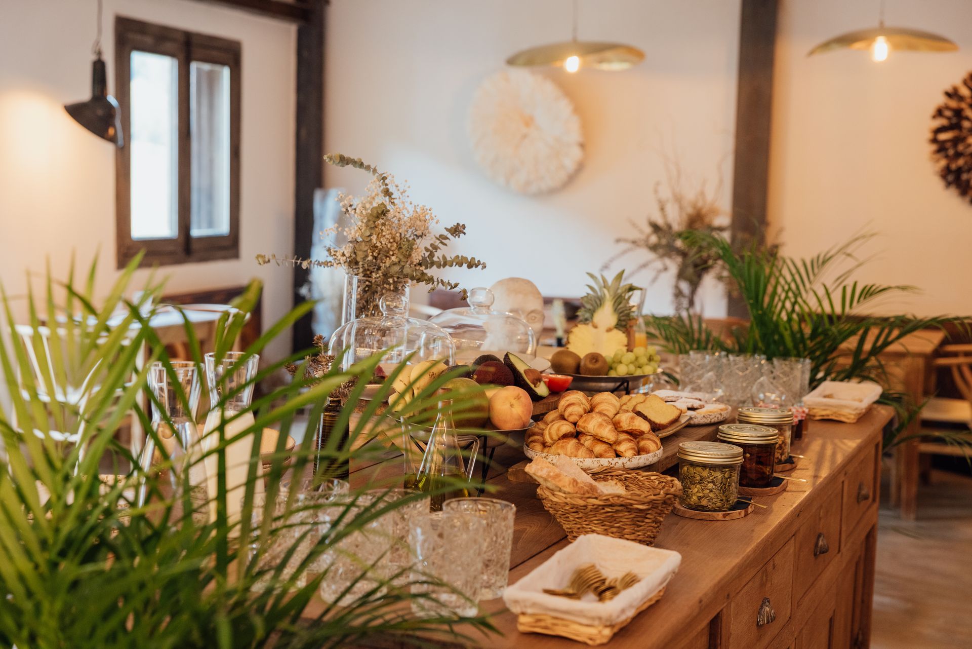 Breakfast buffet with fruit, pastries, and plants on a wooden table.