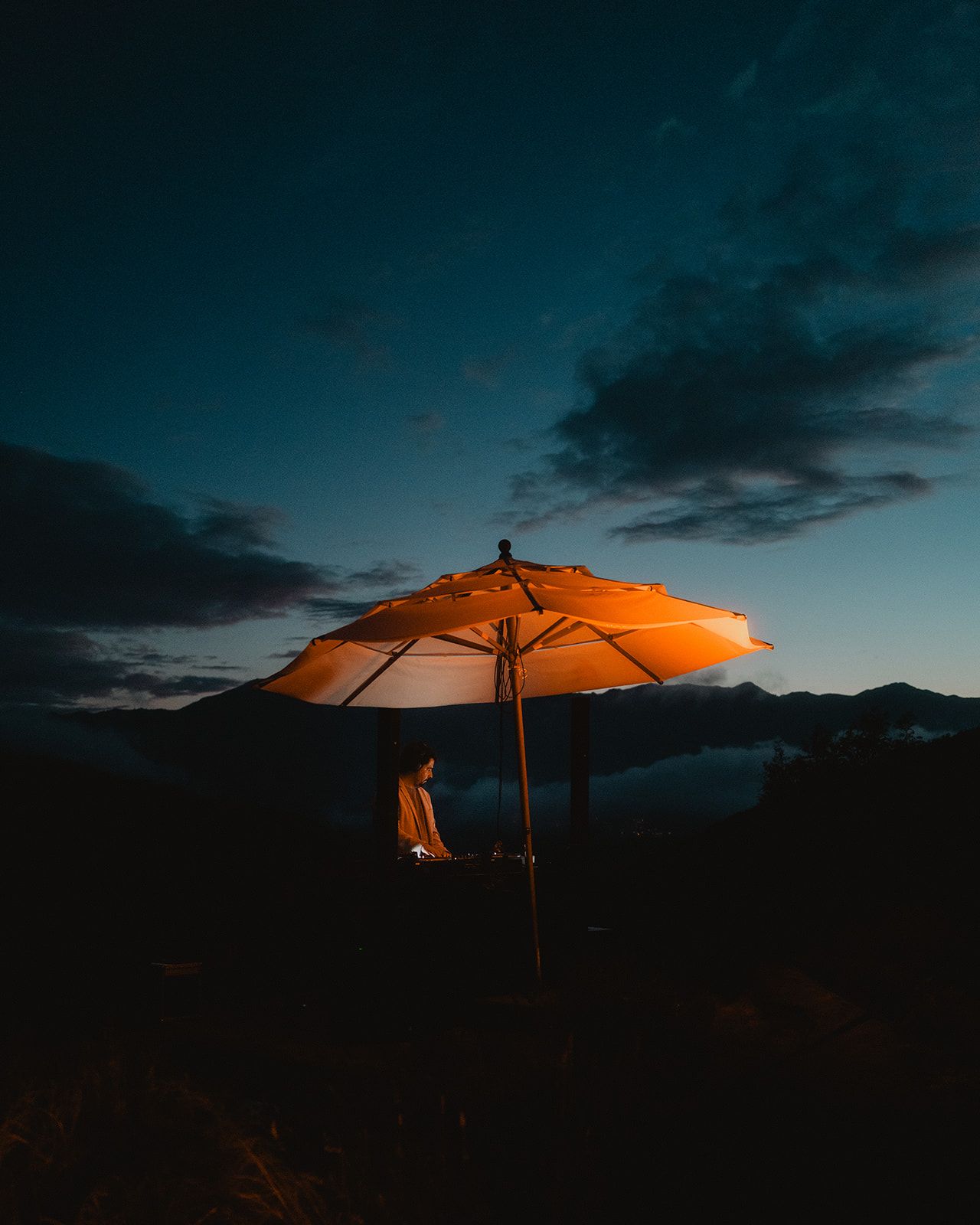 Person under an orange lit umbrella at dusk with mountains in the background.