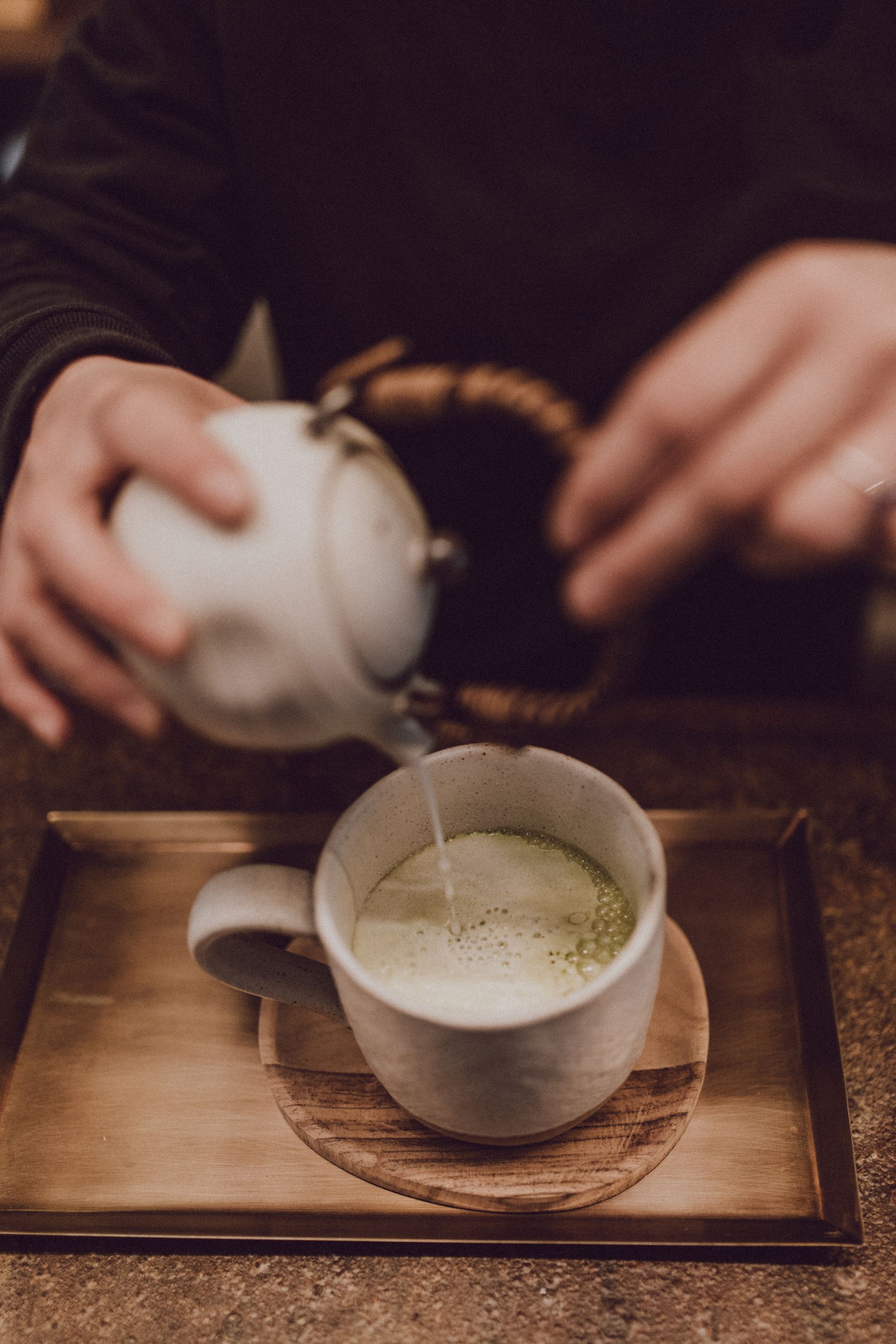Person pours water from a white teapot into a mug of matcha tea on a wooden tray.