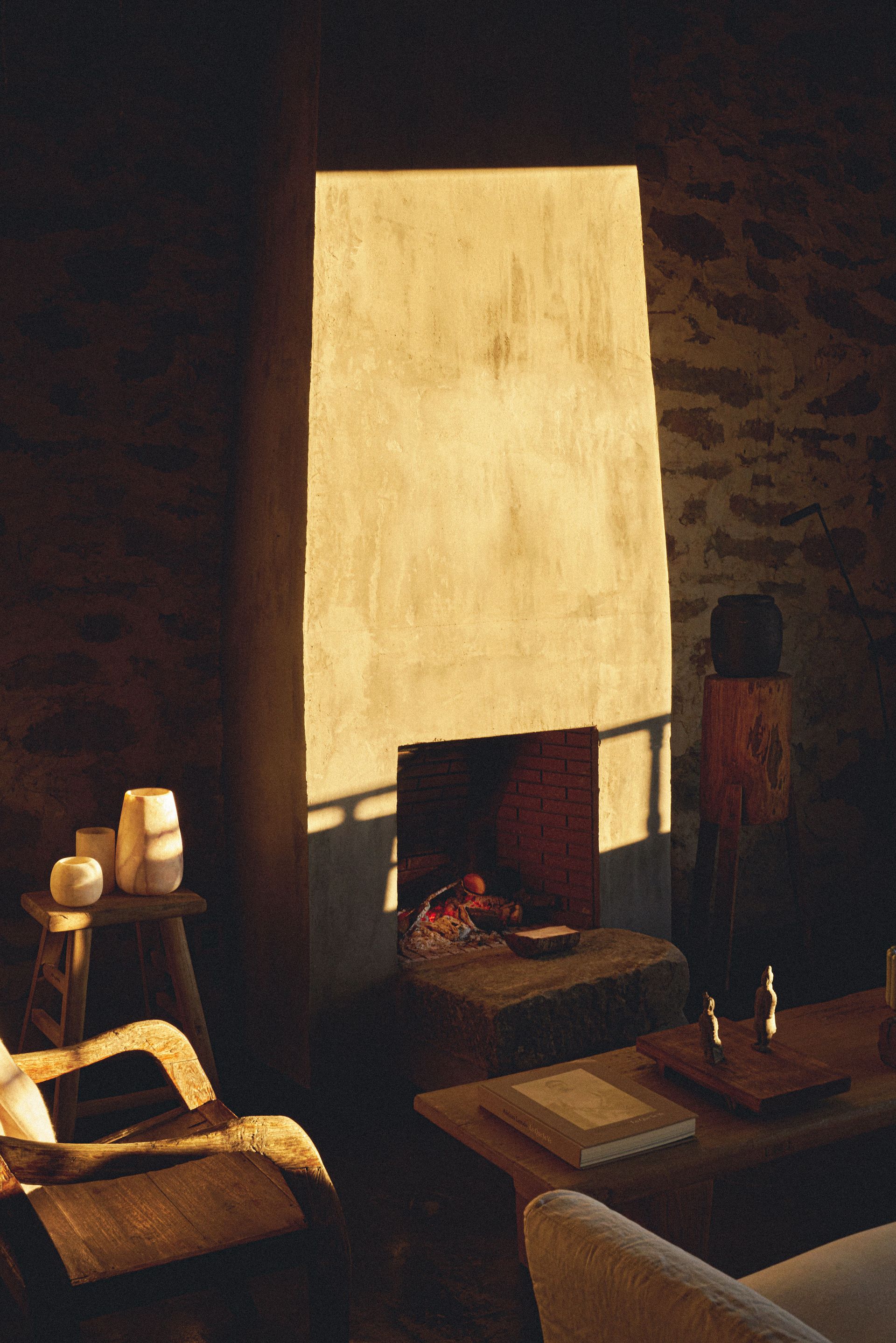 Sunlit fireplace in a stone-walled room; cozy interior with chair, table, and candles.