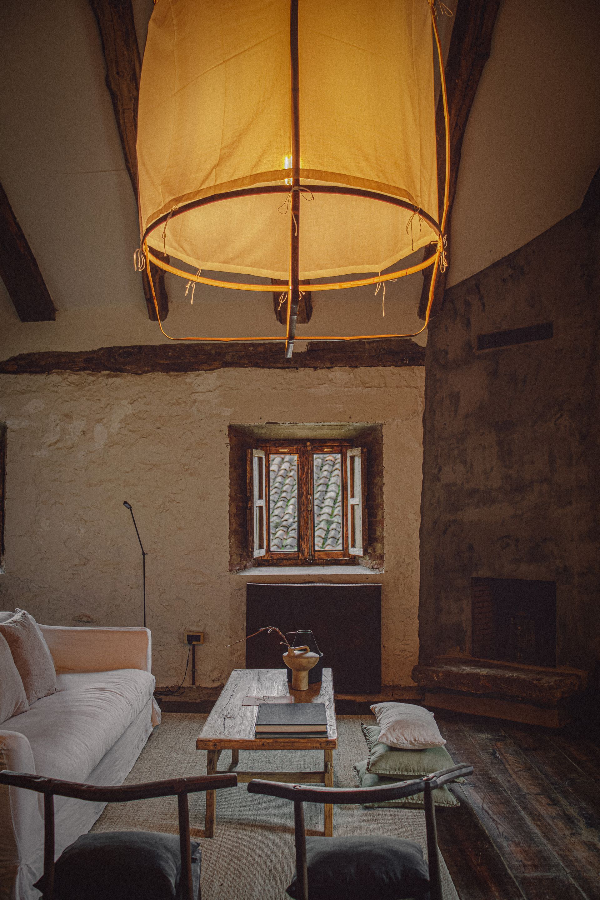 Cozy living room with stone fireplace, window, and large hanging lamp. Light sofa and wooden furniture.