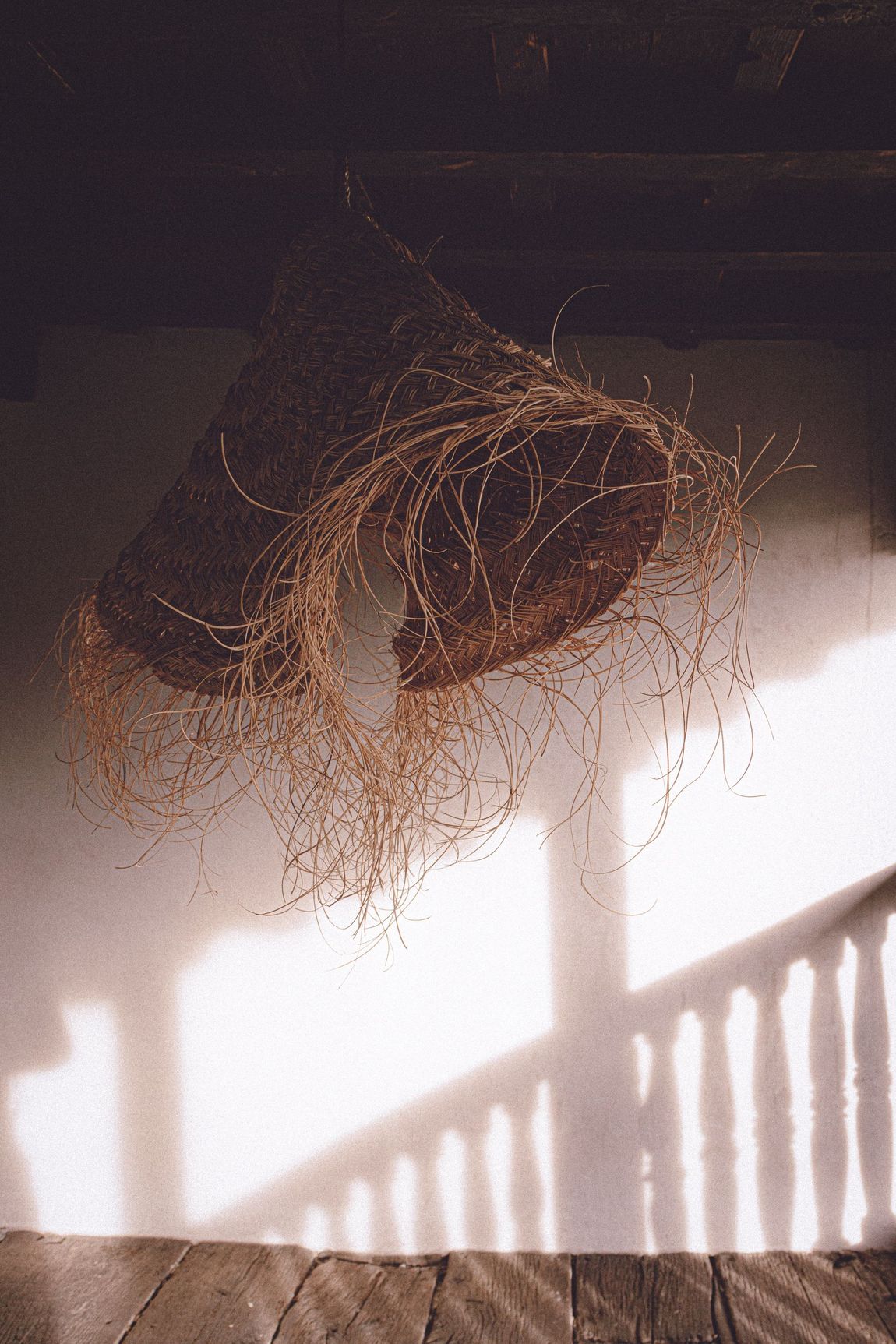Woven brown basket-like structure hanging on a white wall; sunlit shadows from a railing are visible.