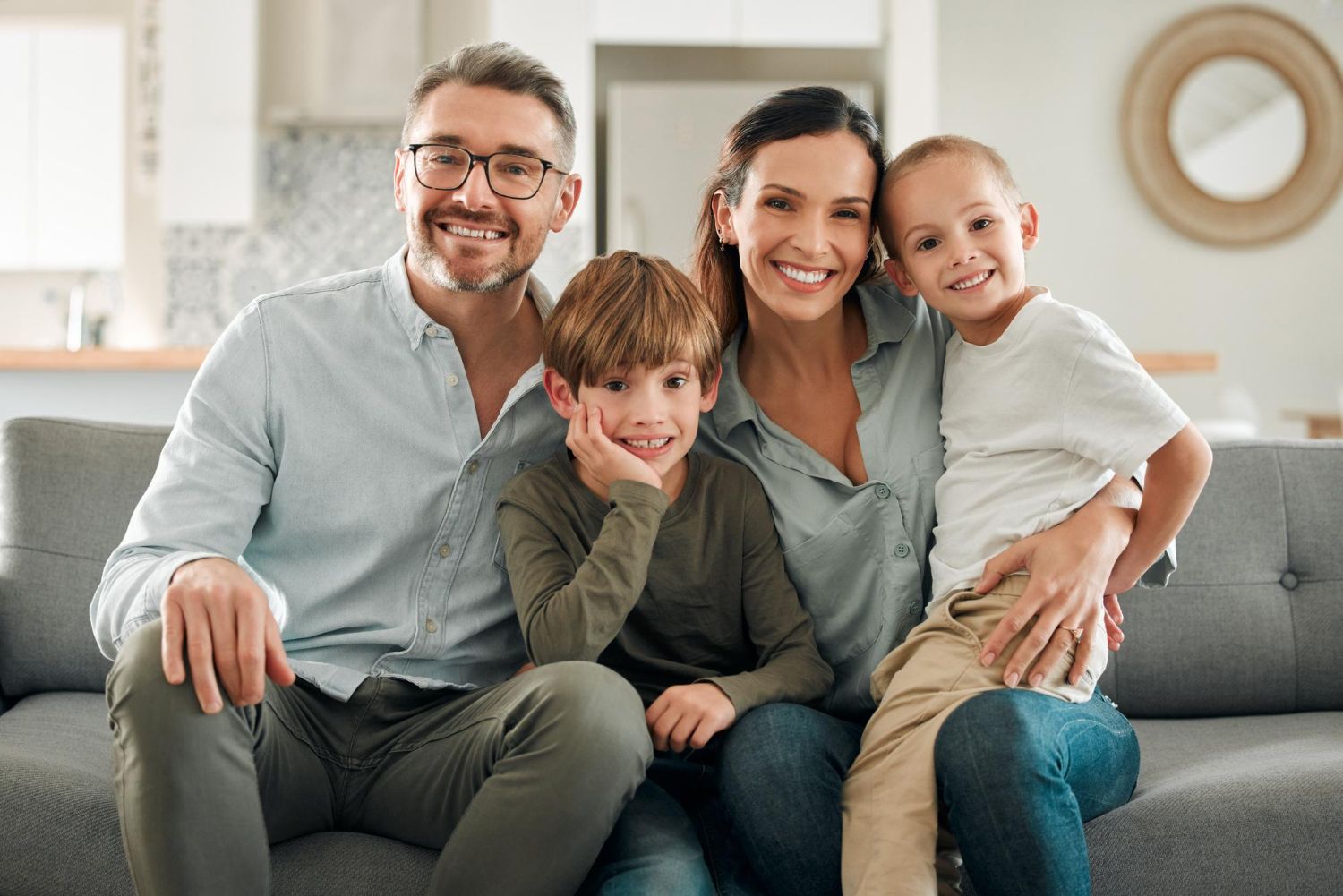 Family of four sitting on a gray couch, smiling in a bright living room.