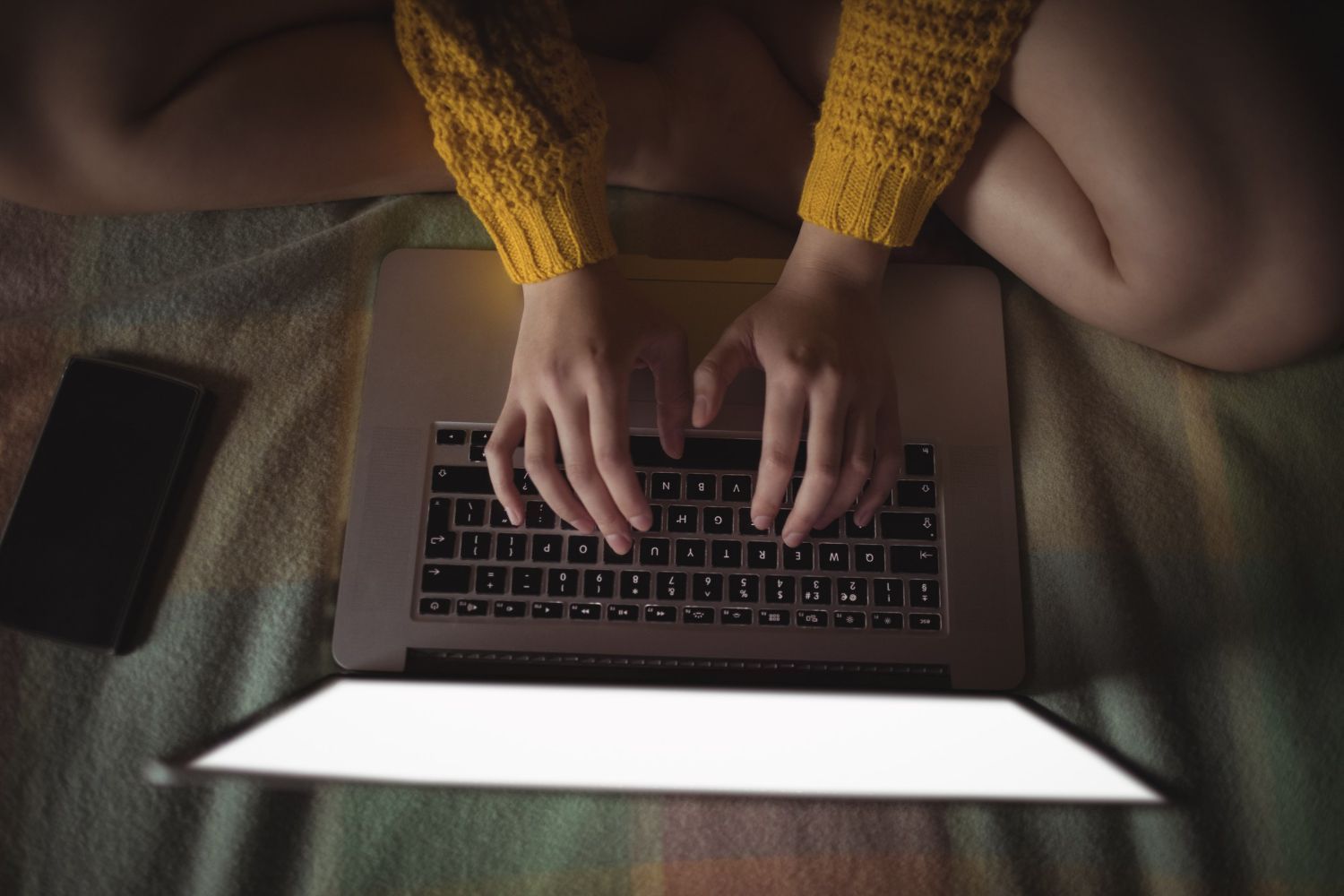 Hands typing on a laptop on a bed, with a bright screen and yellow sleeves visible.
