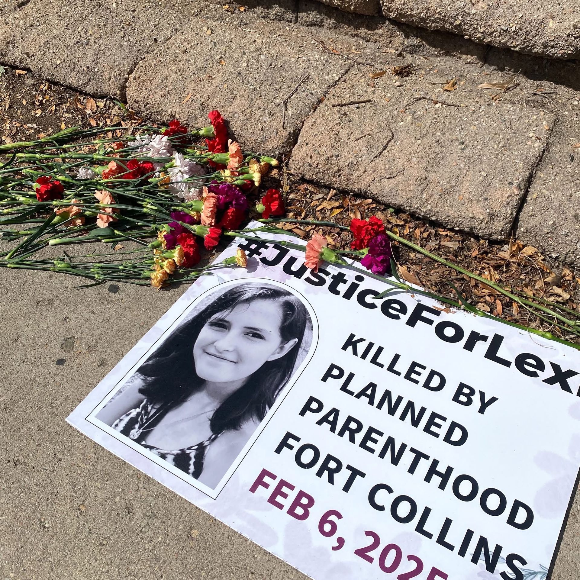 Red, white, pink and yellow carnations scattered on concrete beside brick steps. A placard rests among them bearing a photograph of Lexi Arguello and the words: Hashtag Justice For Lexi. Killed by Planned Parenthood Fort Collins, February 6, 2025.