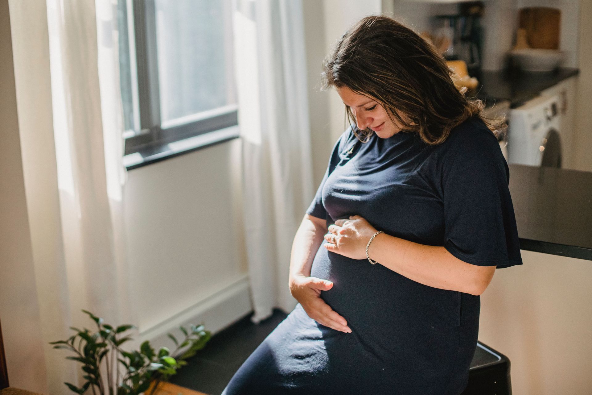 Pregnant person sitting by a window, gently touching their belly in soft daylight