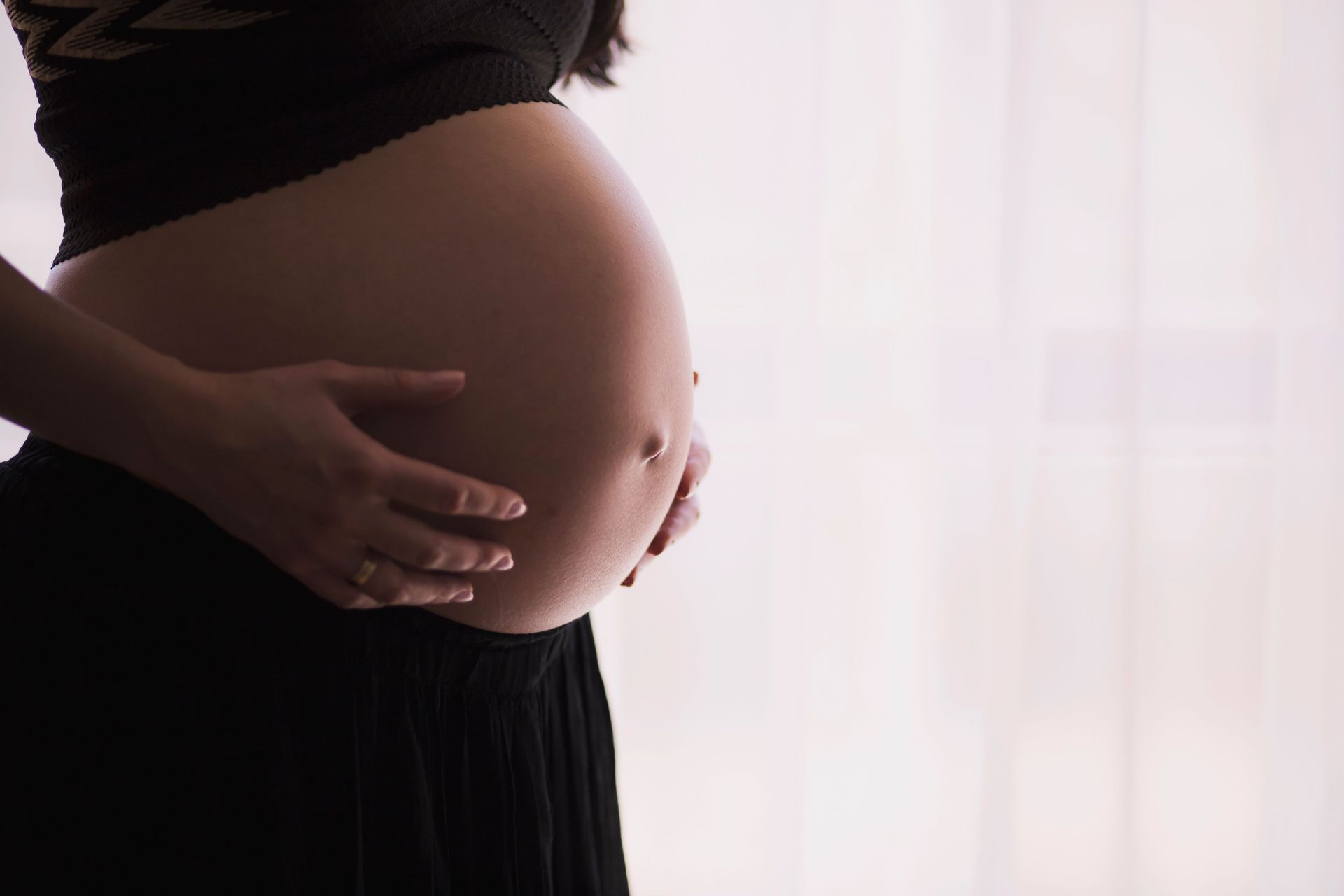 Pregnant belly with hands gently resting on it, backlit by a soft window glow
