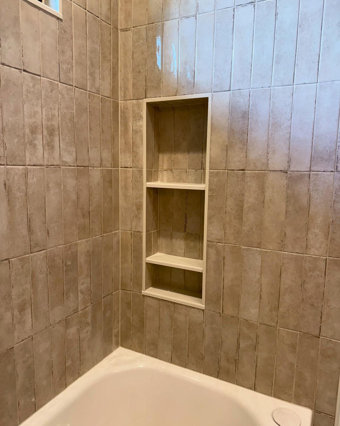 Tiled shower interior with a built-in alcove with three shelves. Beige tiles and white tub.
