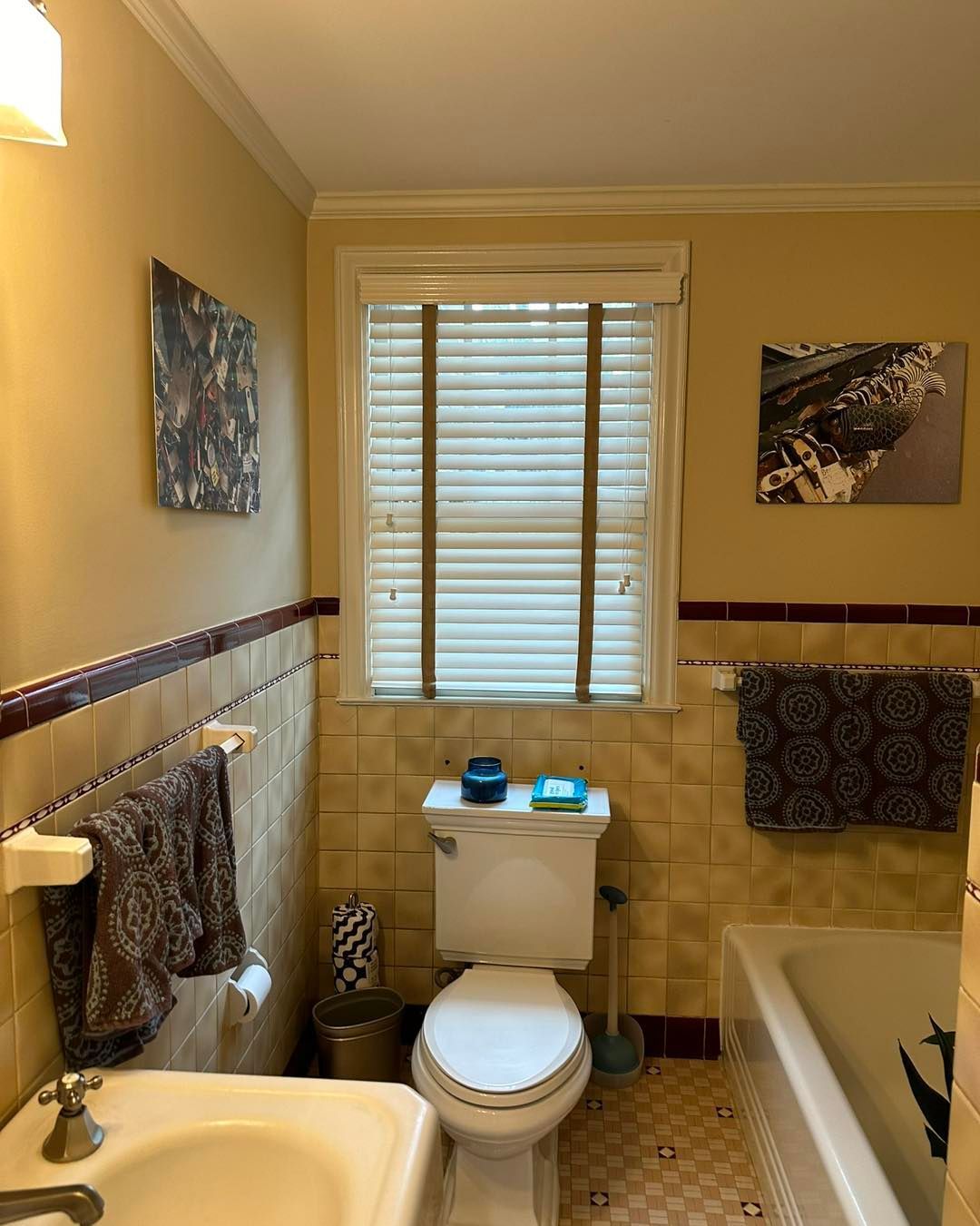 Bathroom with a toilet, window with blinds, and towels on racks. Light yellow walls and beige tile.