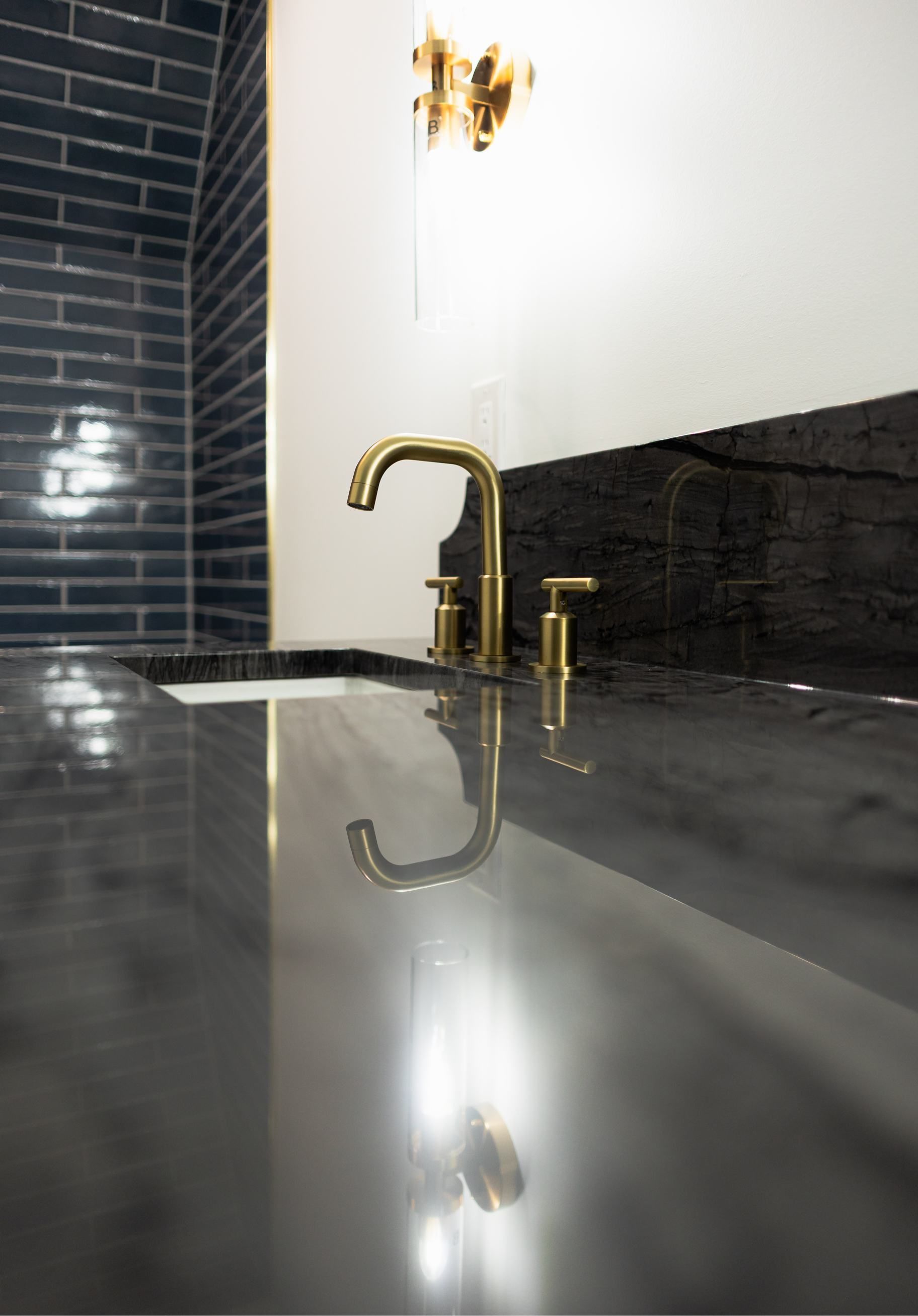 Modern bathroom sink with gold faucet, dark tile backsplash, and glossy gray countertop.